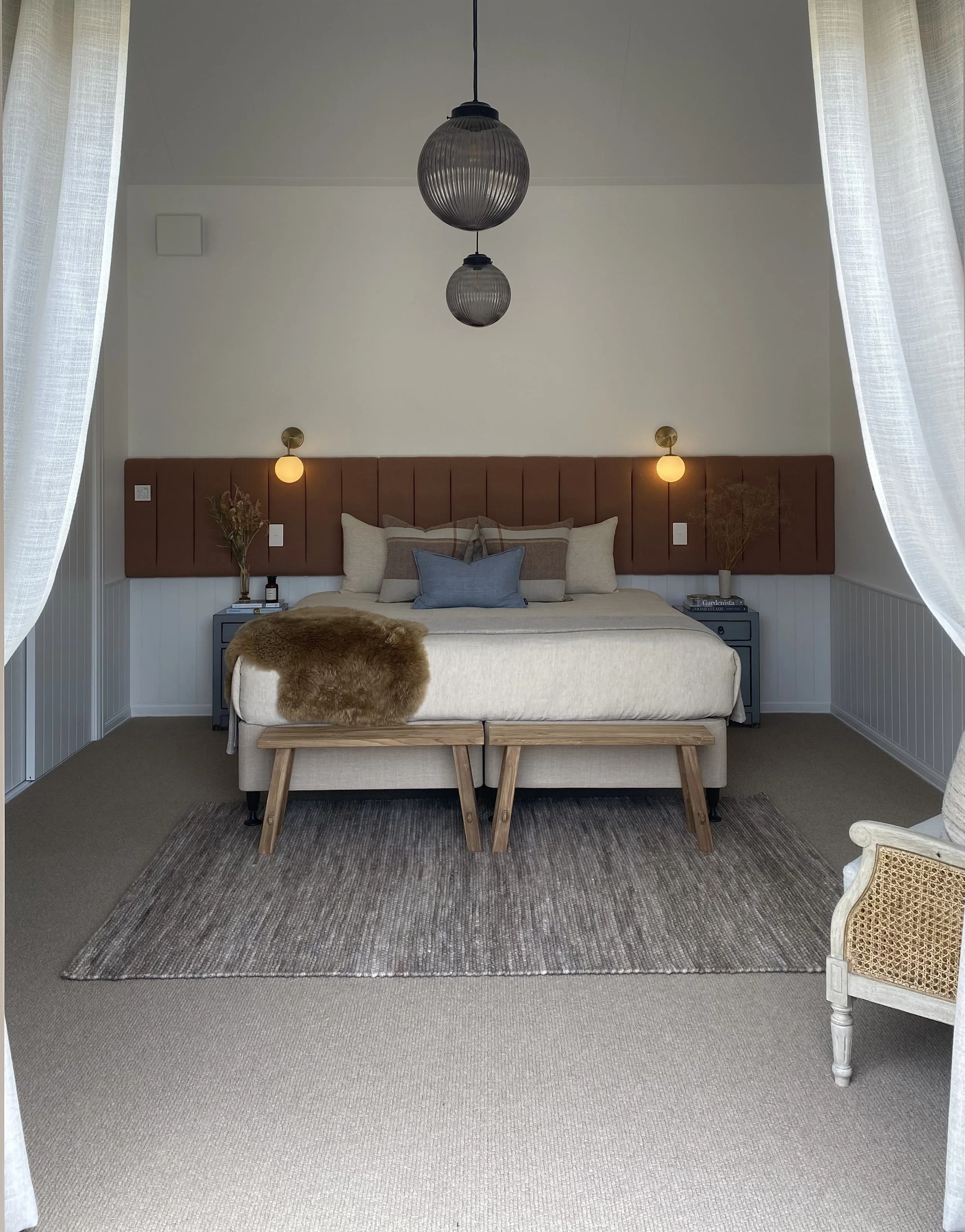 Modern bedroom with beige bed, brown headboard, two blue nightstands, bedside lamps, and a beige rug. Curtains frame the scene, and black pendant lights hang from the ceiling.
