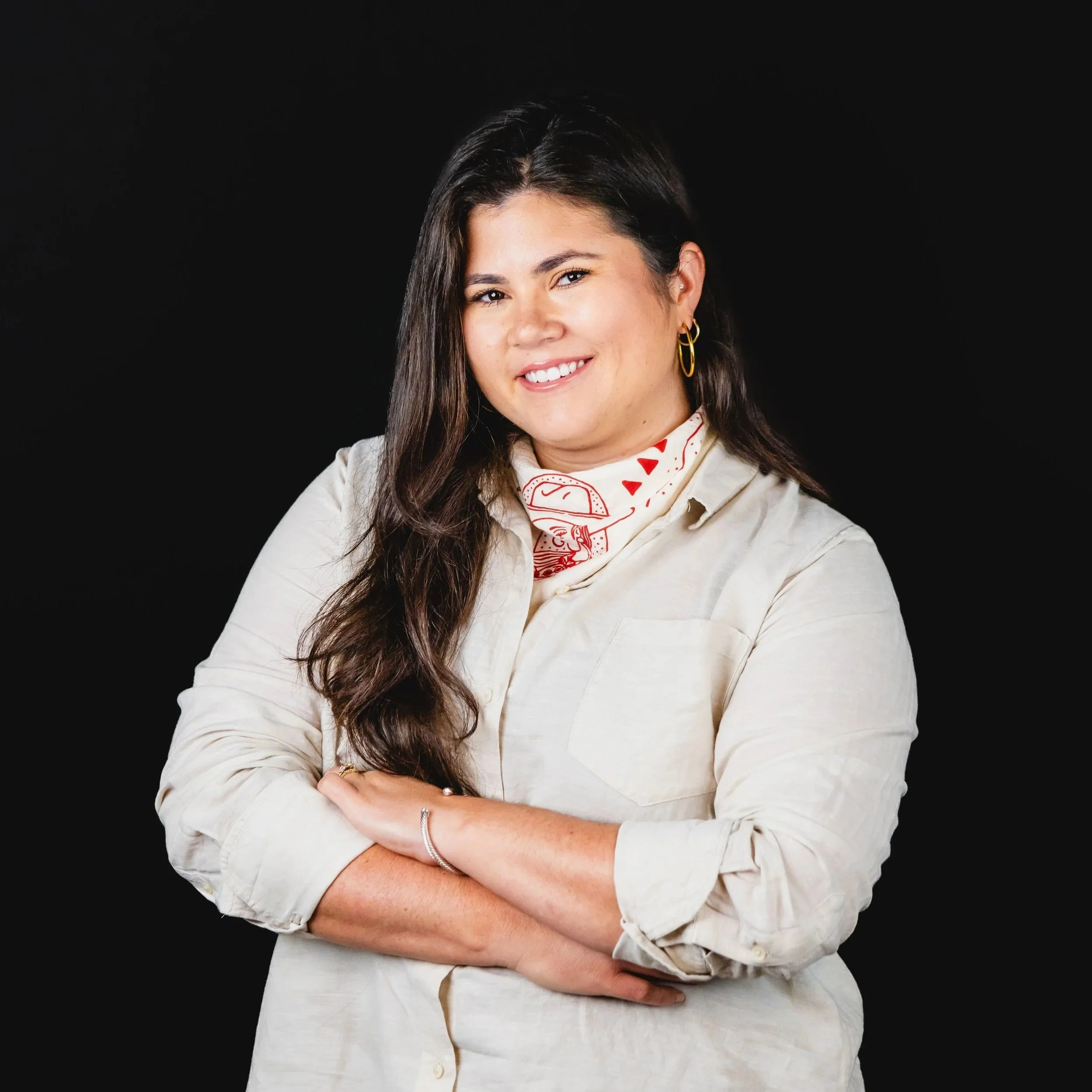 A woman with long dark hair smiling, wearing a cream-colored shirt and a white bandana with red designs, standing against a black background.