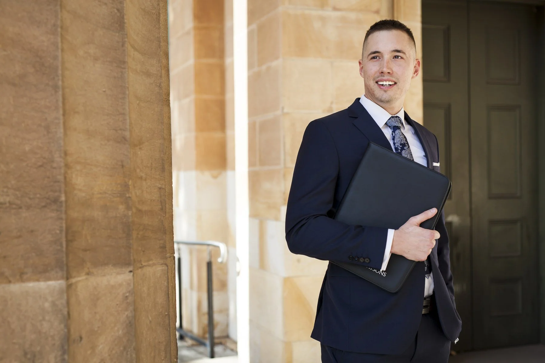 A man in a navy suit holding a black laptop folder, smiling, standing outside a building with sandstone walls and large dark green door.