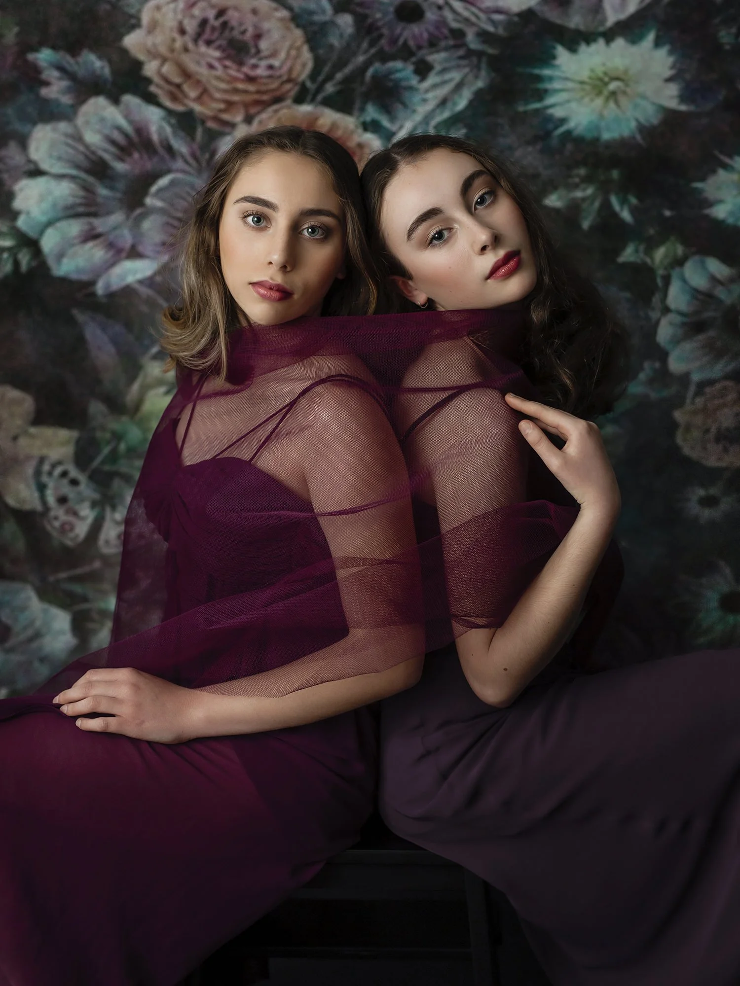 Two women posed together with a floral background, dressed in sheer, dark red fabrics, with their arms around each other and gazing into the camera.