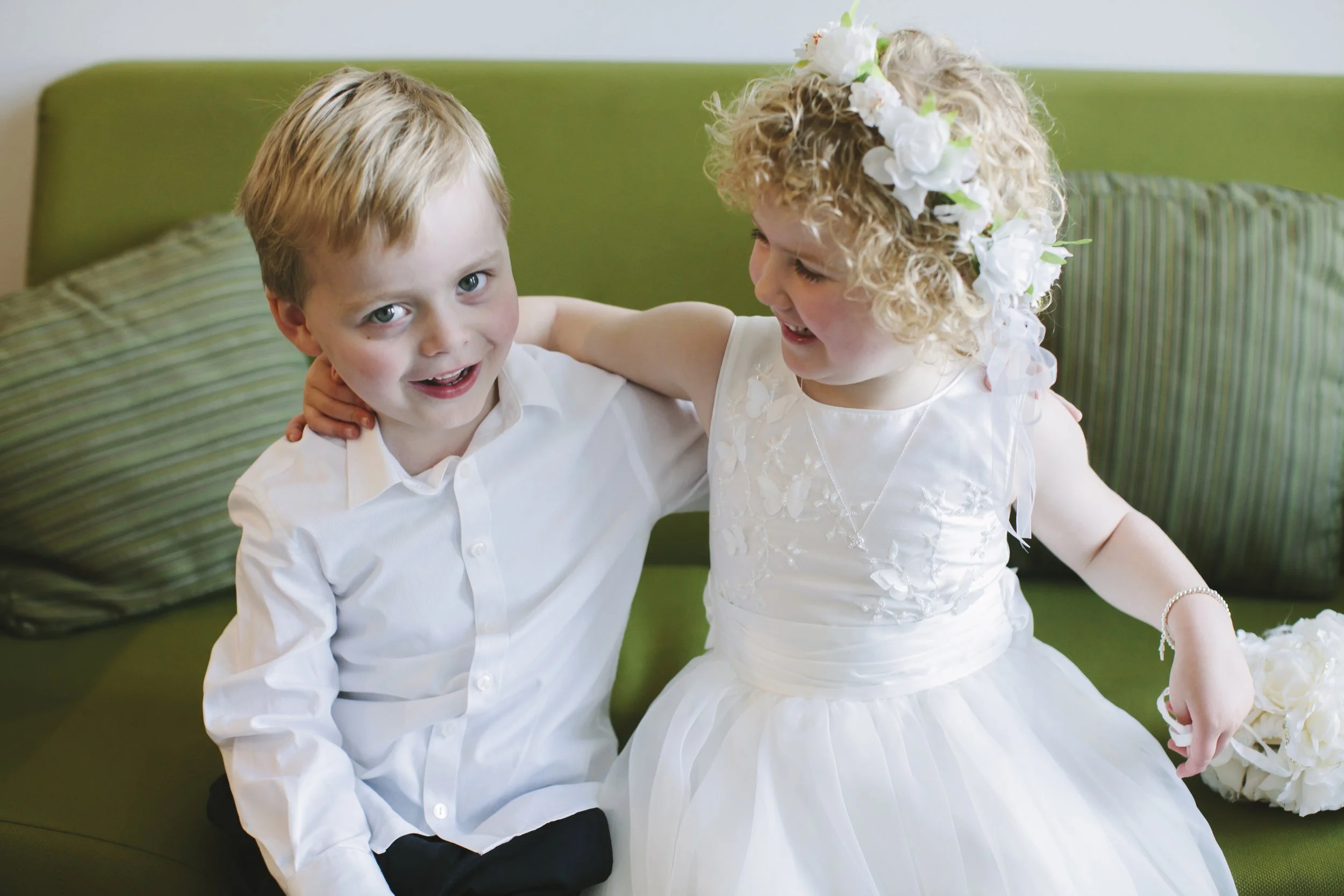 Two young children, a boy and a girl, dressed in wedding attire, sitting on a green sofa with striped pillows, smiling and interacting happily.