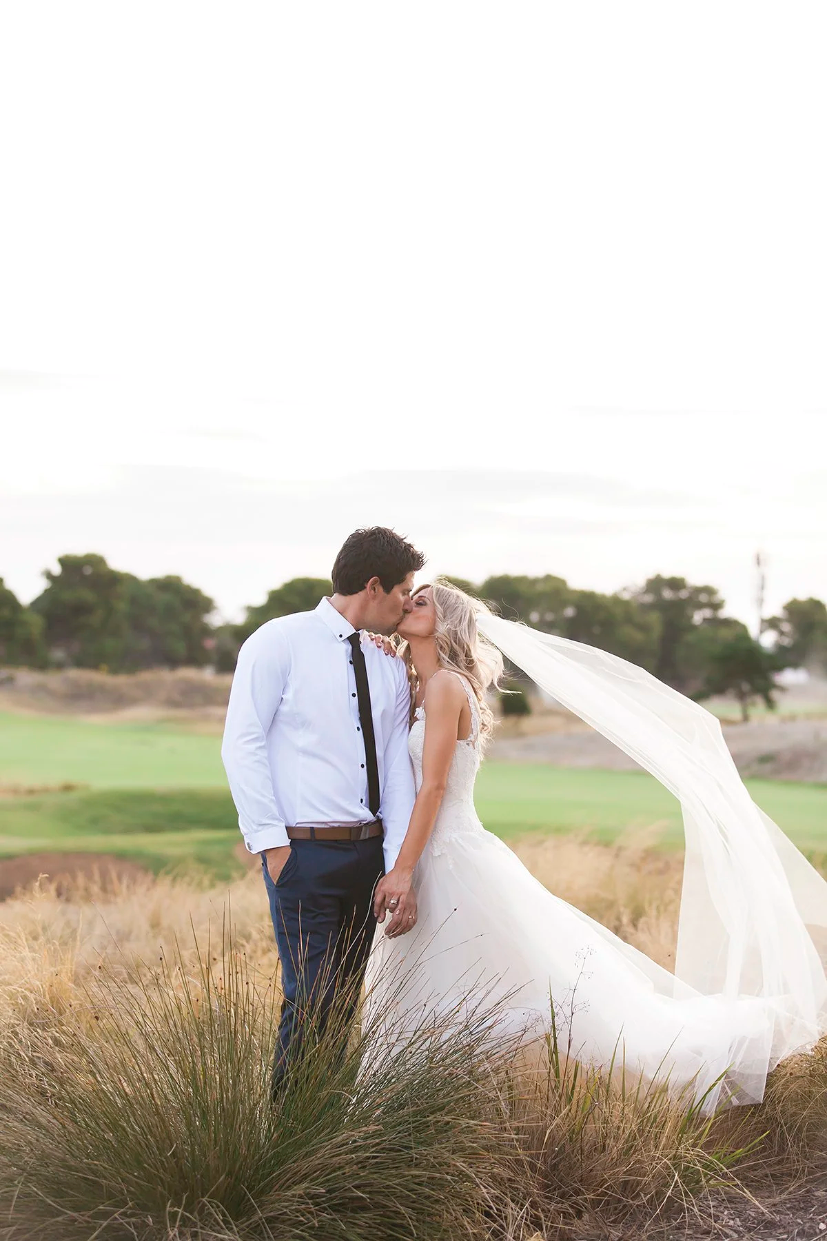 A bride and groom kissing outdoors in a field with dry grass, trees, and cloudy sky in the background.
