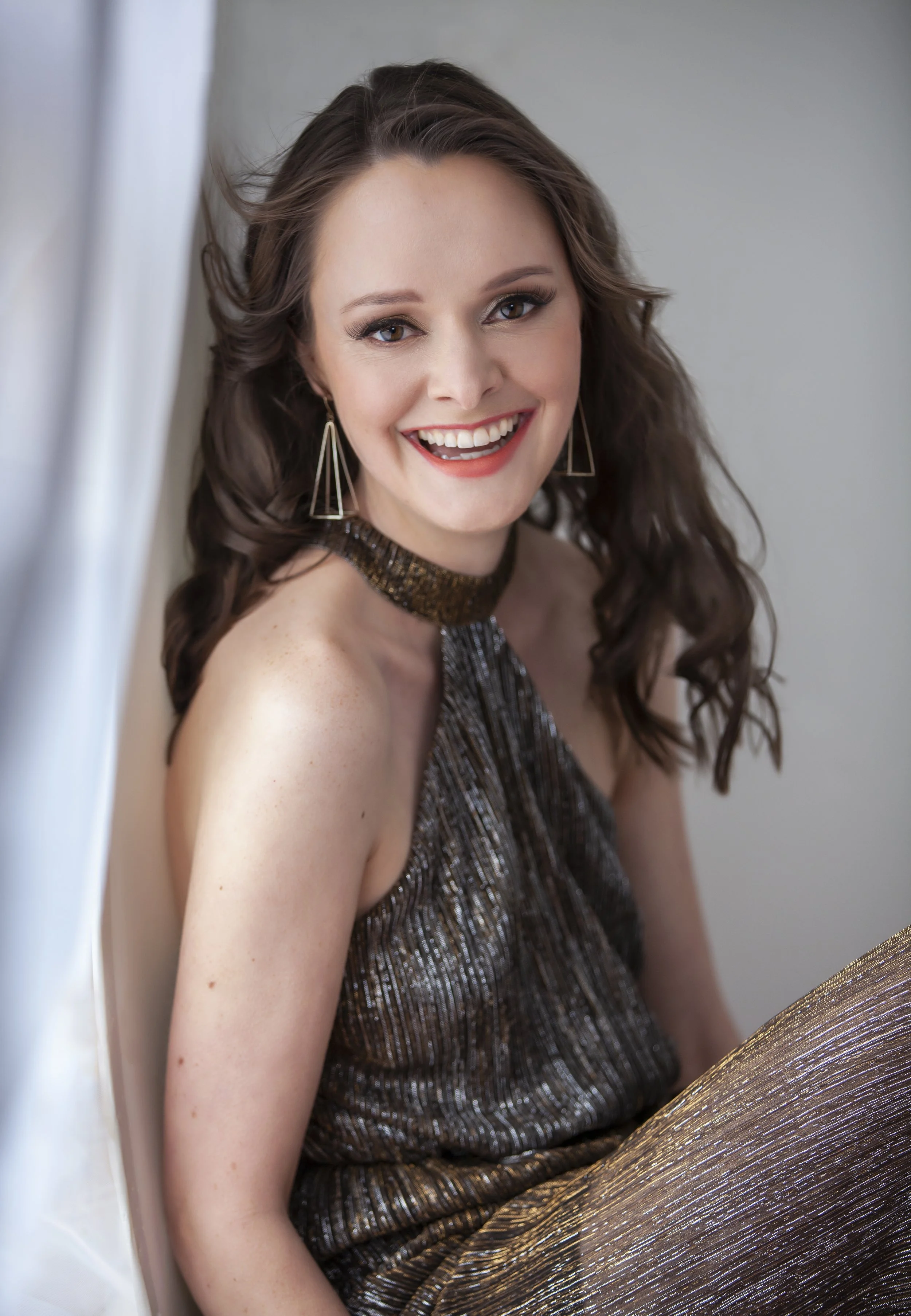A woman with wavy brown hair smiling, wearing a sparkly metallic dress, gold earrings, and a choker, seated near a window with natural light.