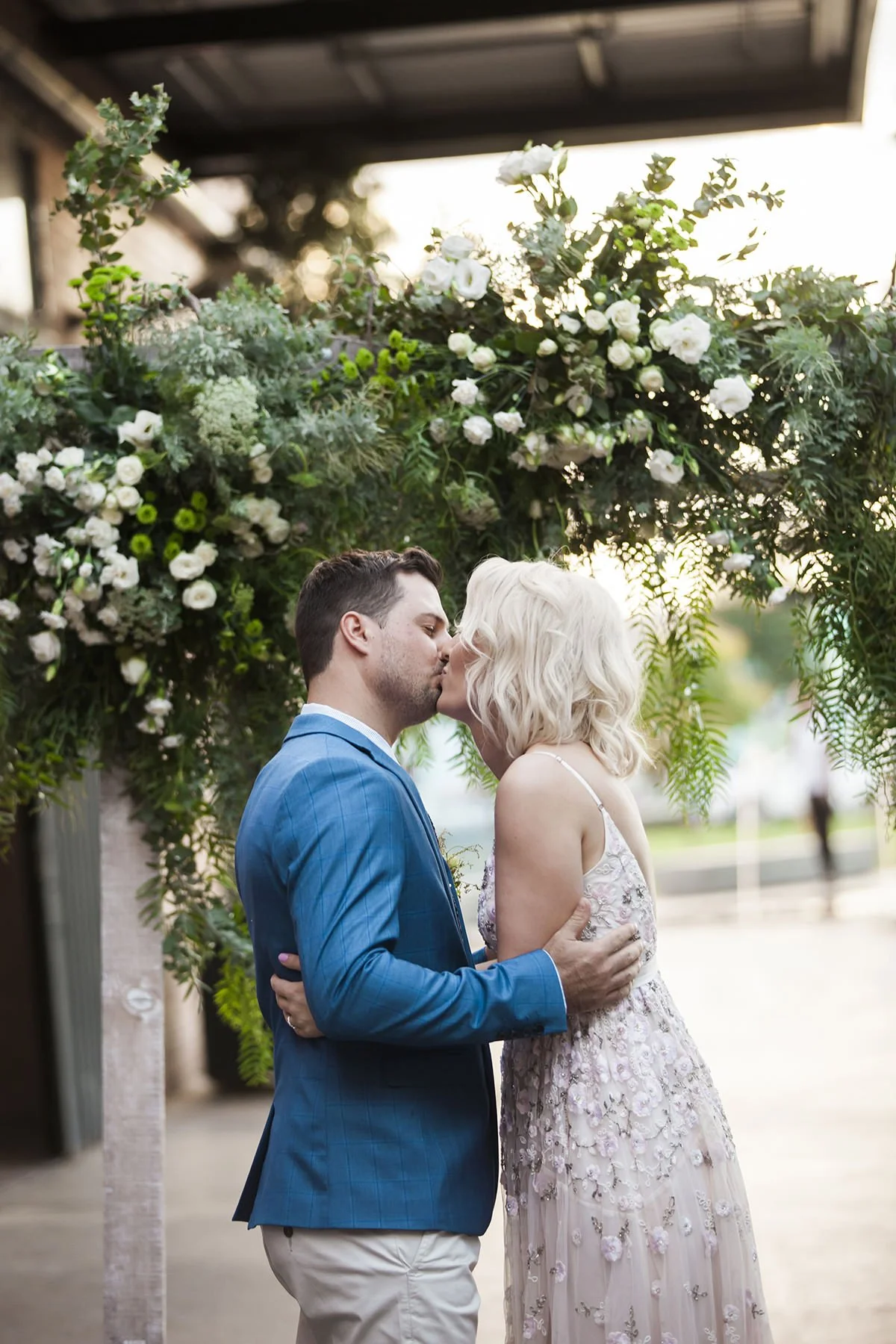 A couple kissing at their wedding under a floral arch, with the groom in a blue suit and the bride in a white lace dress, during sunset.