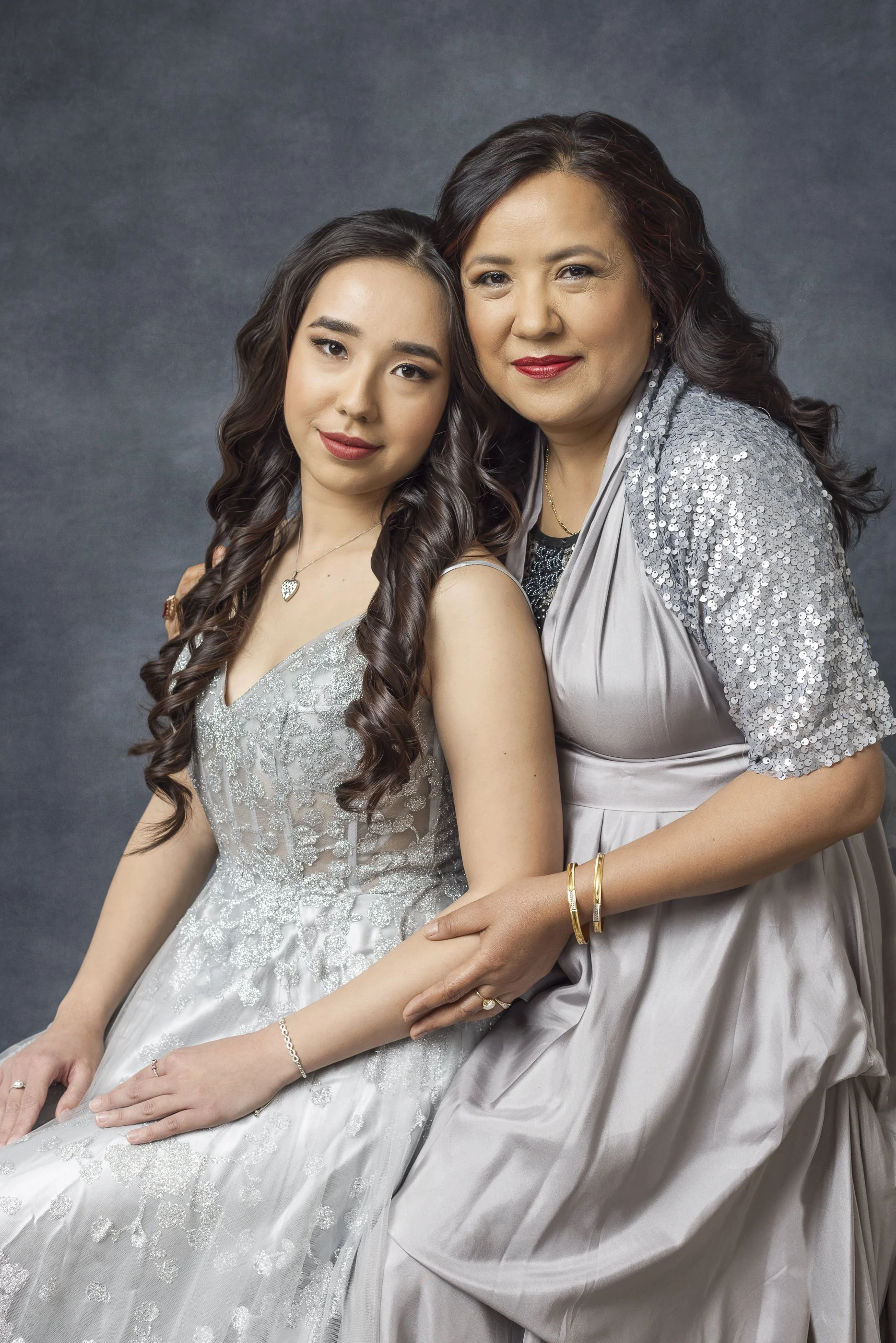 A young woman in a silver dress with lace details sitting next to an older woman in a matching silver outfit with sequined sleeves, both smiling and posing together against a gray background.