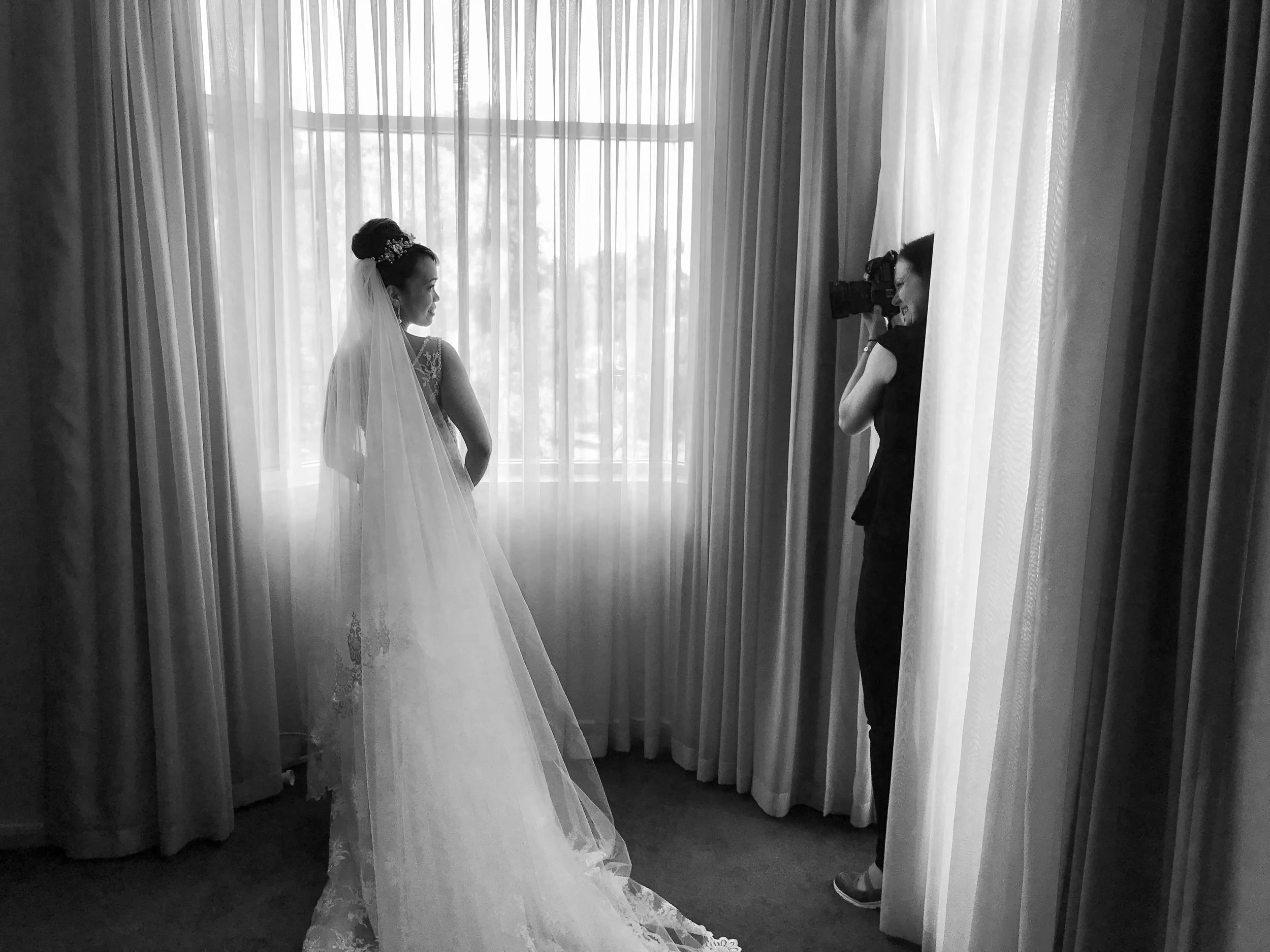 A bride in a wedding gown and veil stands near a window while a photographer captures her photo inside a room with curtains.