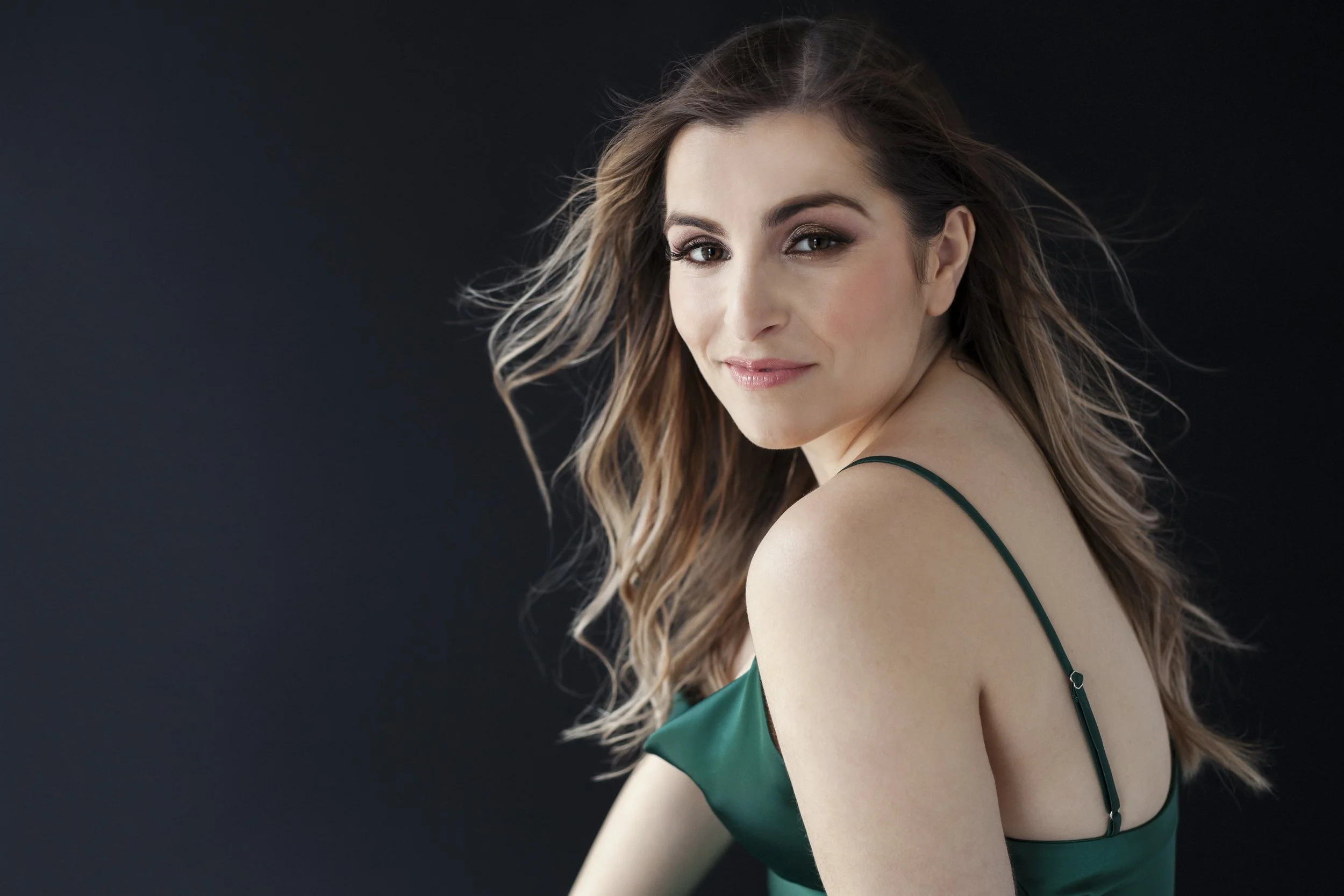 A woman with long, wavy brown hair and fair skin wearing a green satin dress with spaghetti straps, against a dark background.