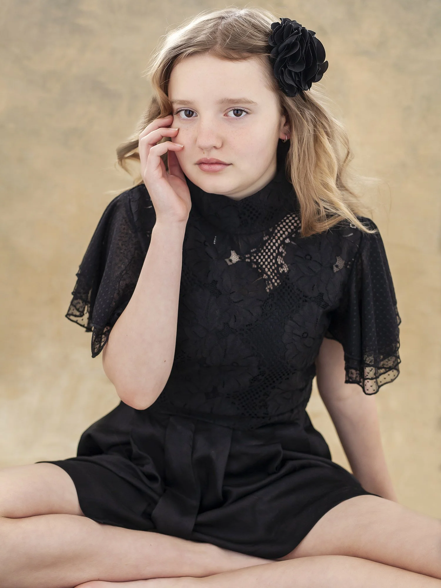 A young girl with wavy blonde hair, wearing a black floral lace top and matching shorts, sitting with legs crossed against a beige background, with a black flower hair accessory on the side of her head.