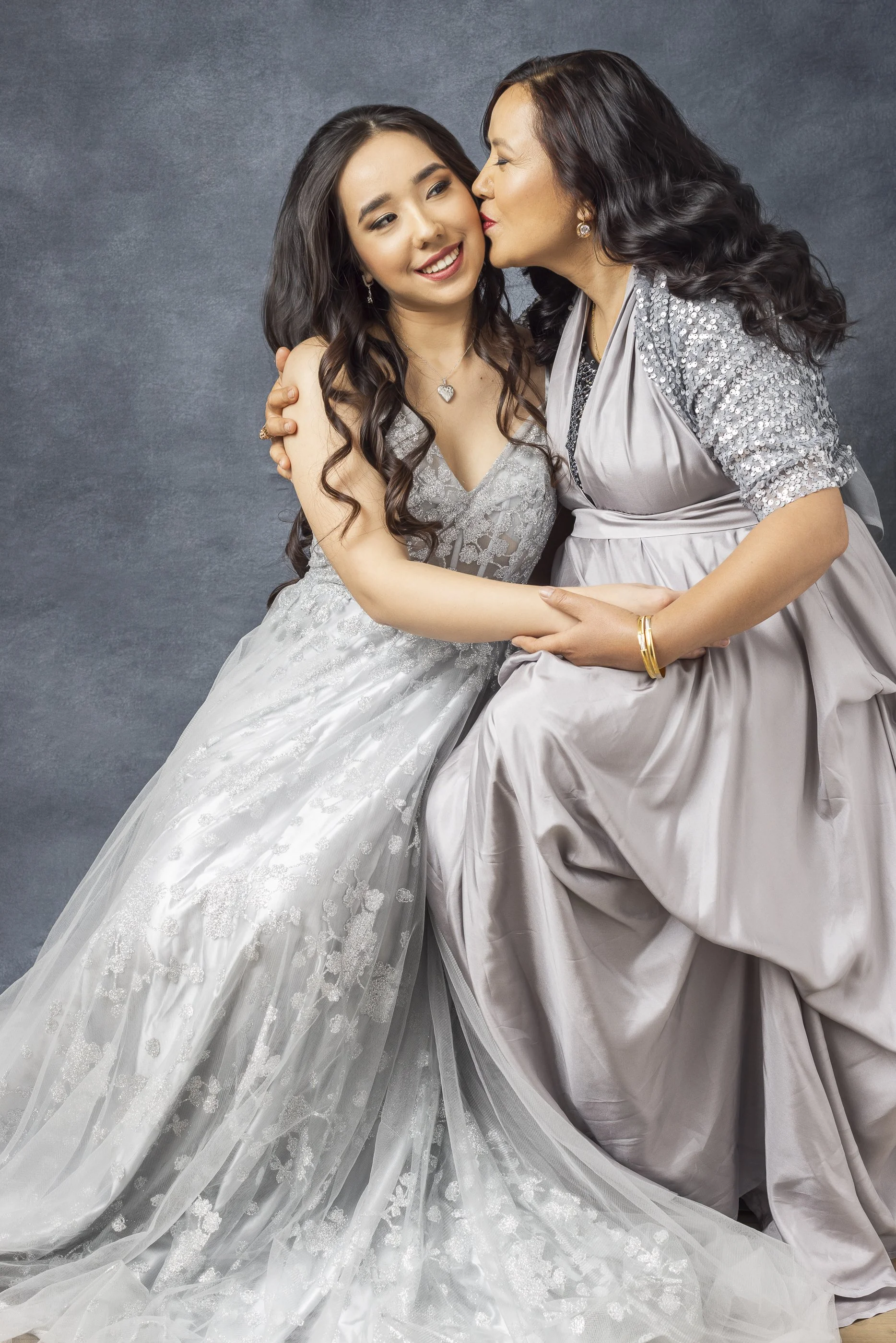 A young woman in a wedding dress being kissed on the cheek by another woman wearing a silver gown.