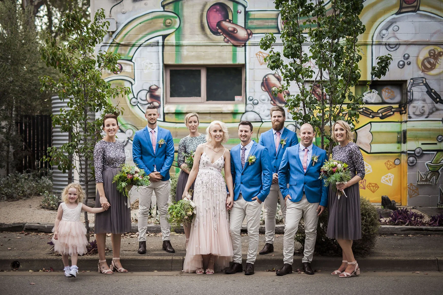 A group of people, including a bride in a pink dress and seven others in blue and gray attire, standing outdoors in front of a colorful graffiti wall with trees and plants.
