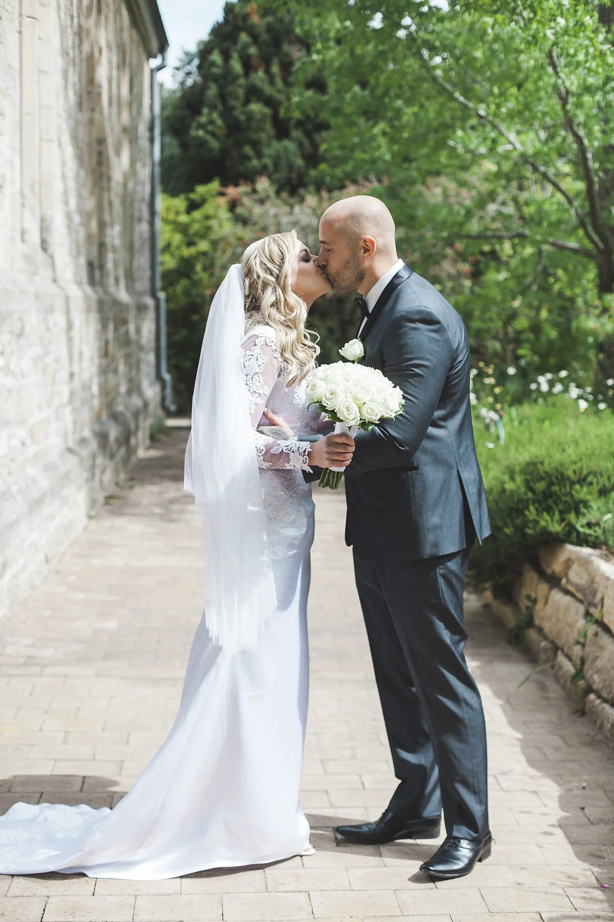 A bride and groom share a kiss outdoors on a brick pathway, surrounded by green trees. The bride wears a white wedding dress and veil, holding a bouquet of white roses. The groom wears a dark suit and tie.