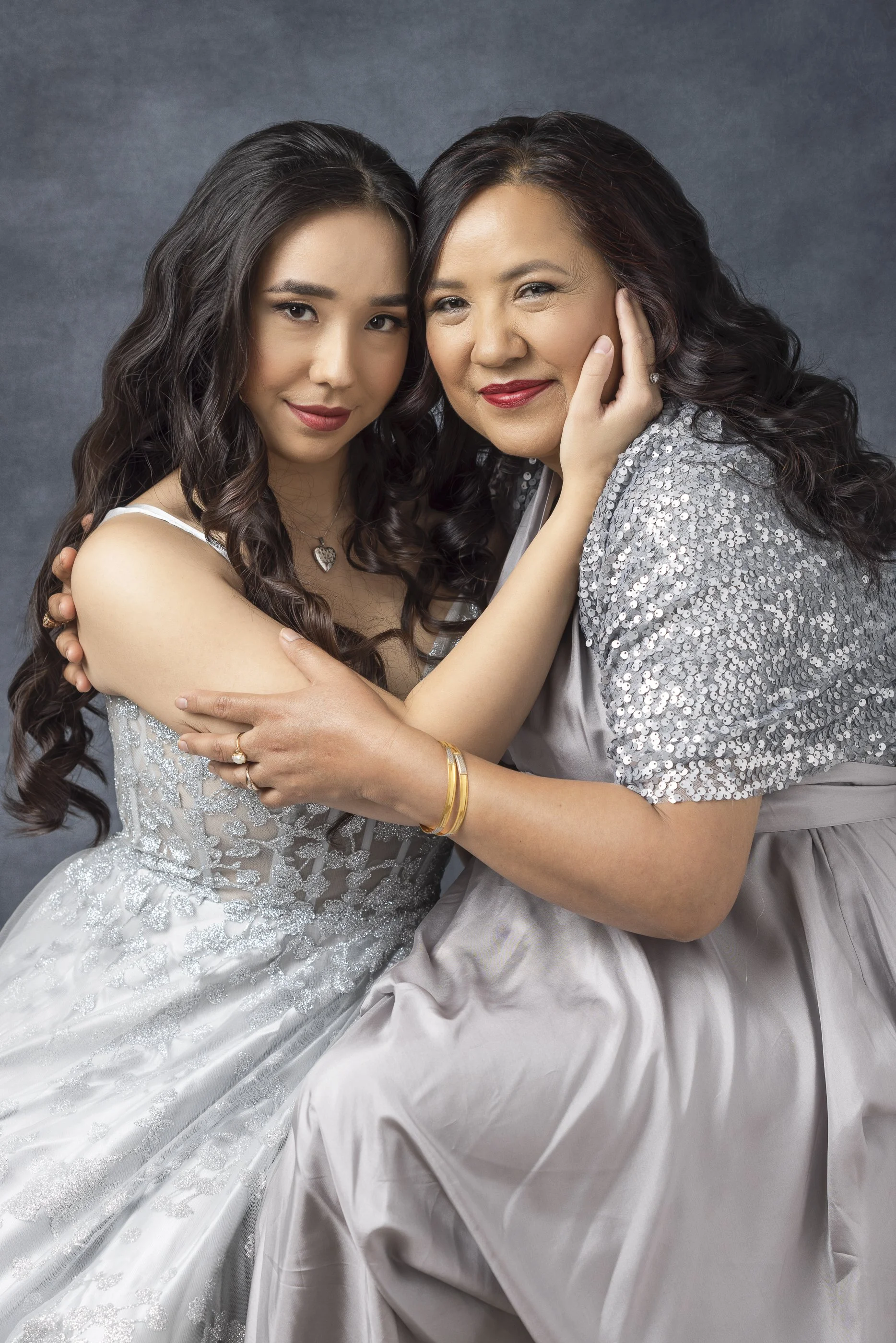 A young woman in a wedding dress and an older woman in a gray dress with sequins are embracing and smiling at the camera against a gray background.