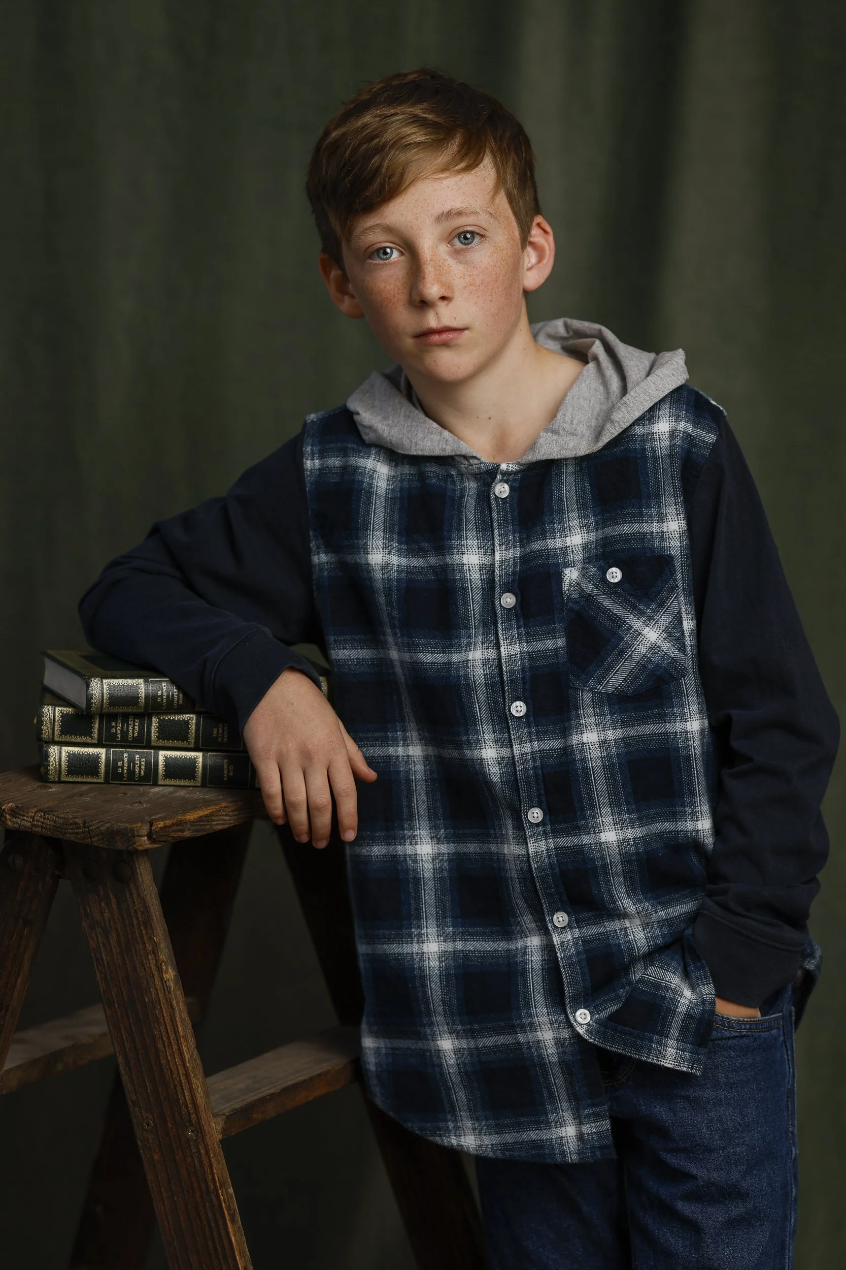 A young boy with red hair, blue eyes, and freckles wearing a blue and black plaid shirt with a gray hood, leaning on a wooden stool with his left arm resting on it, and standing with his right hand in his pocket against a dark green backdrop.