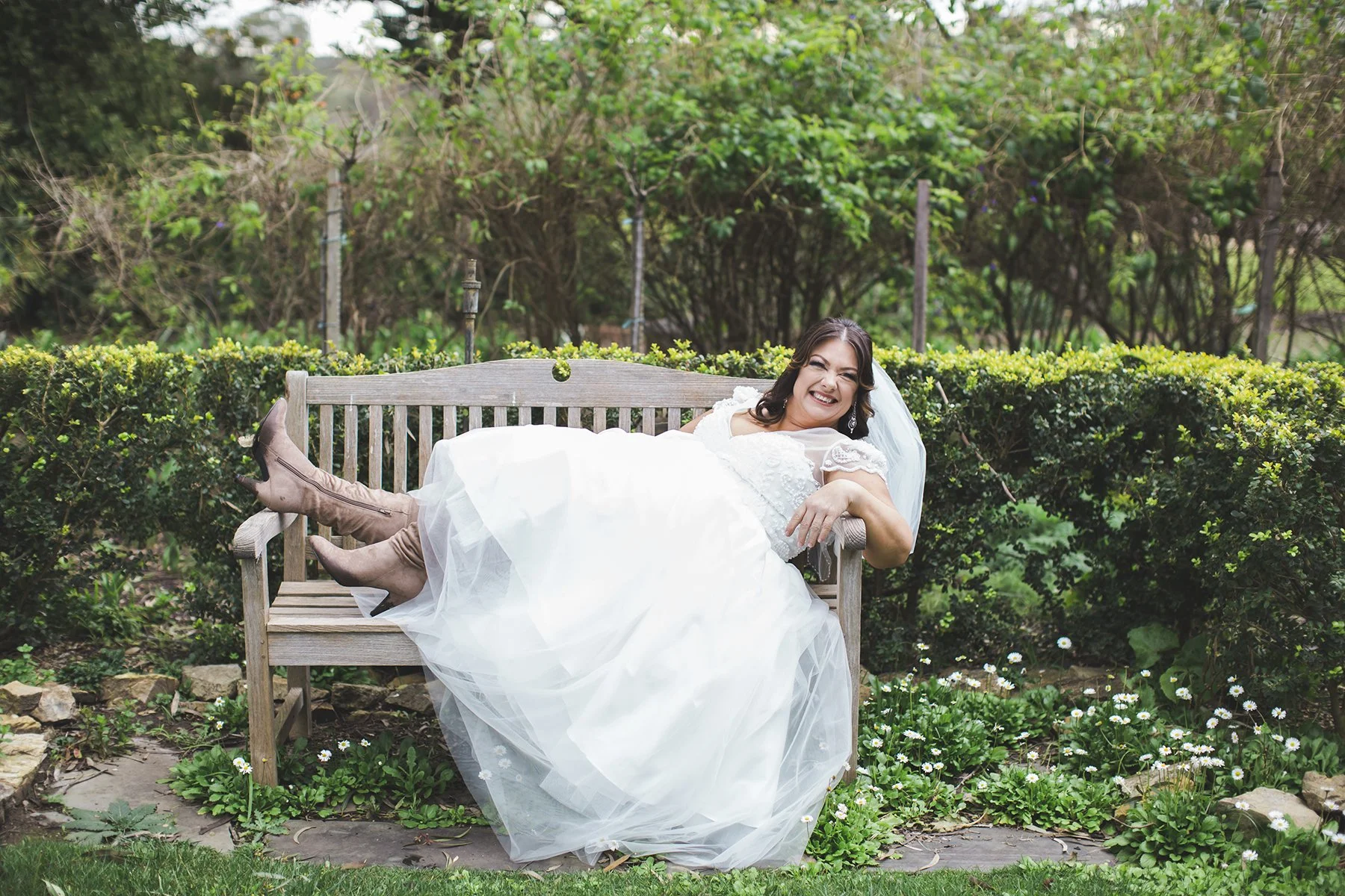 A woman in a wedding dress and brown boots is lying on a wooden garden bench, smiling happily, in a lush green garden with flowers.