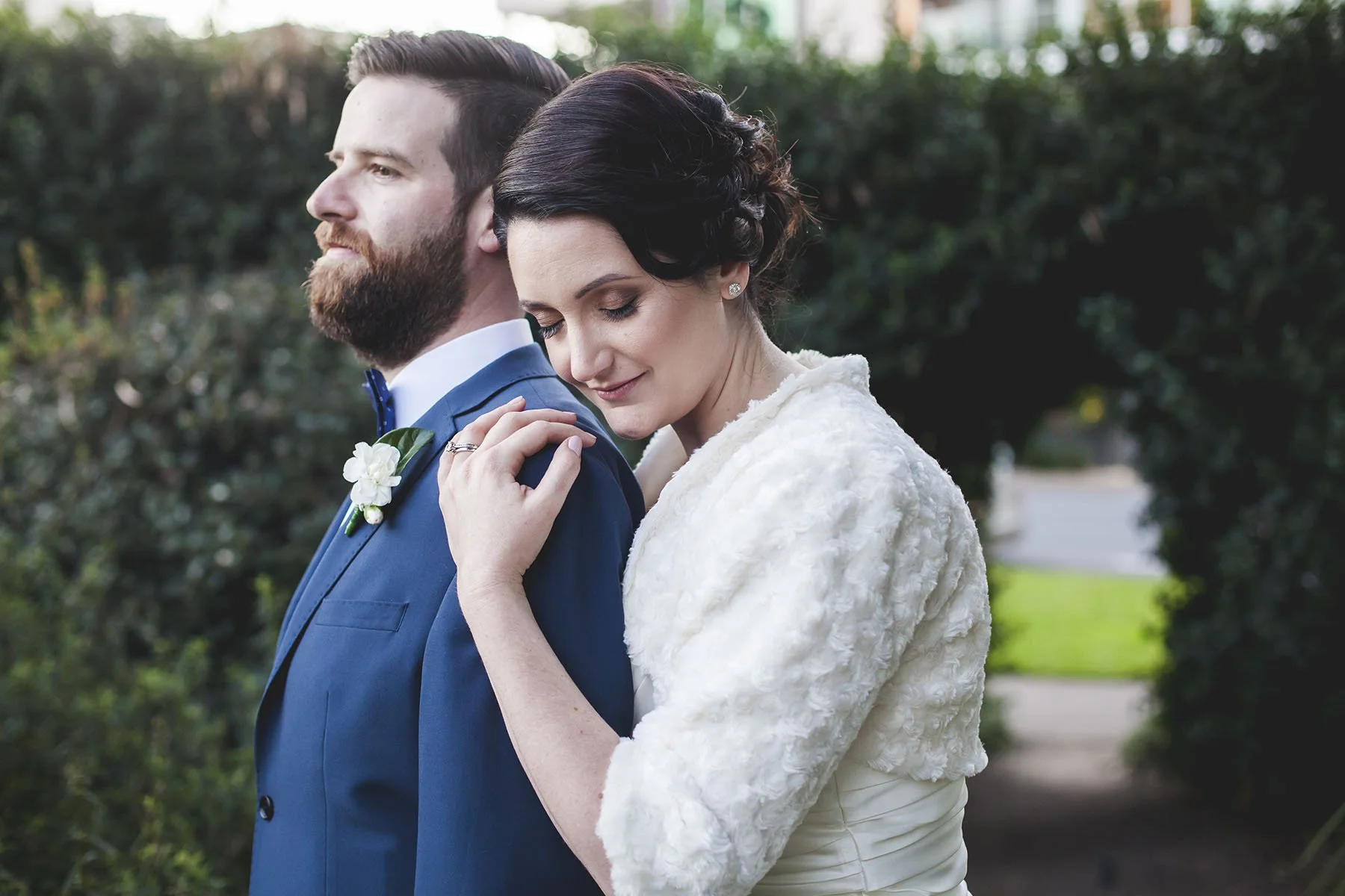 A bride and groom standing close together outdoors on a wedding day, the woman resting her head on the man's shoulder with her eyes closed, wearing a white dress and a textured white jacket, and the man in a blue suit with a white shirt and boutonnie