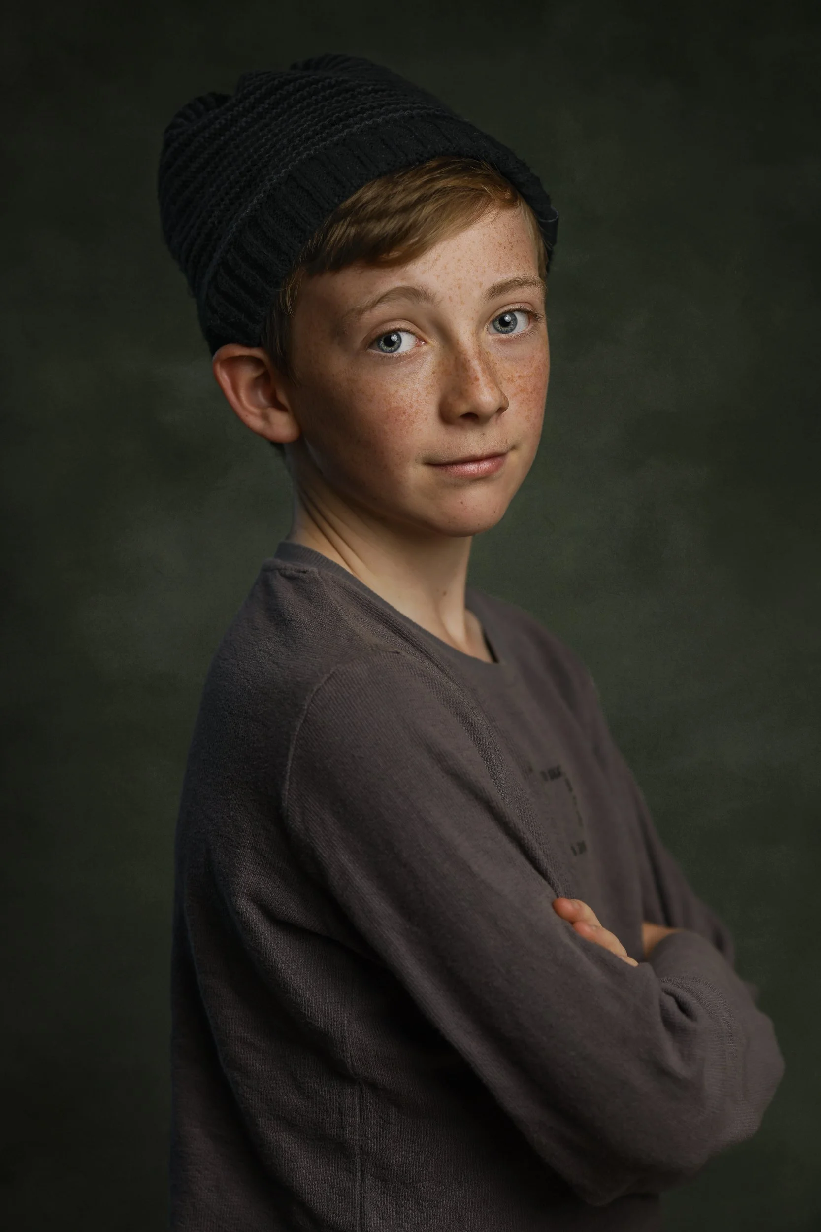 Portrait of a young boy with red hair, freckles, and blue eyes, wearing a black knit beanie and a dark gray sweatshirt, standing with arms crossed against a dark green background.