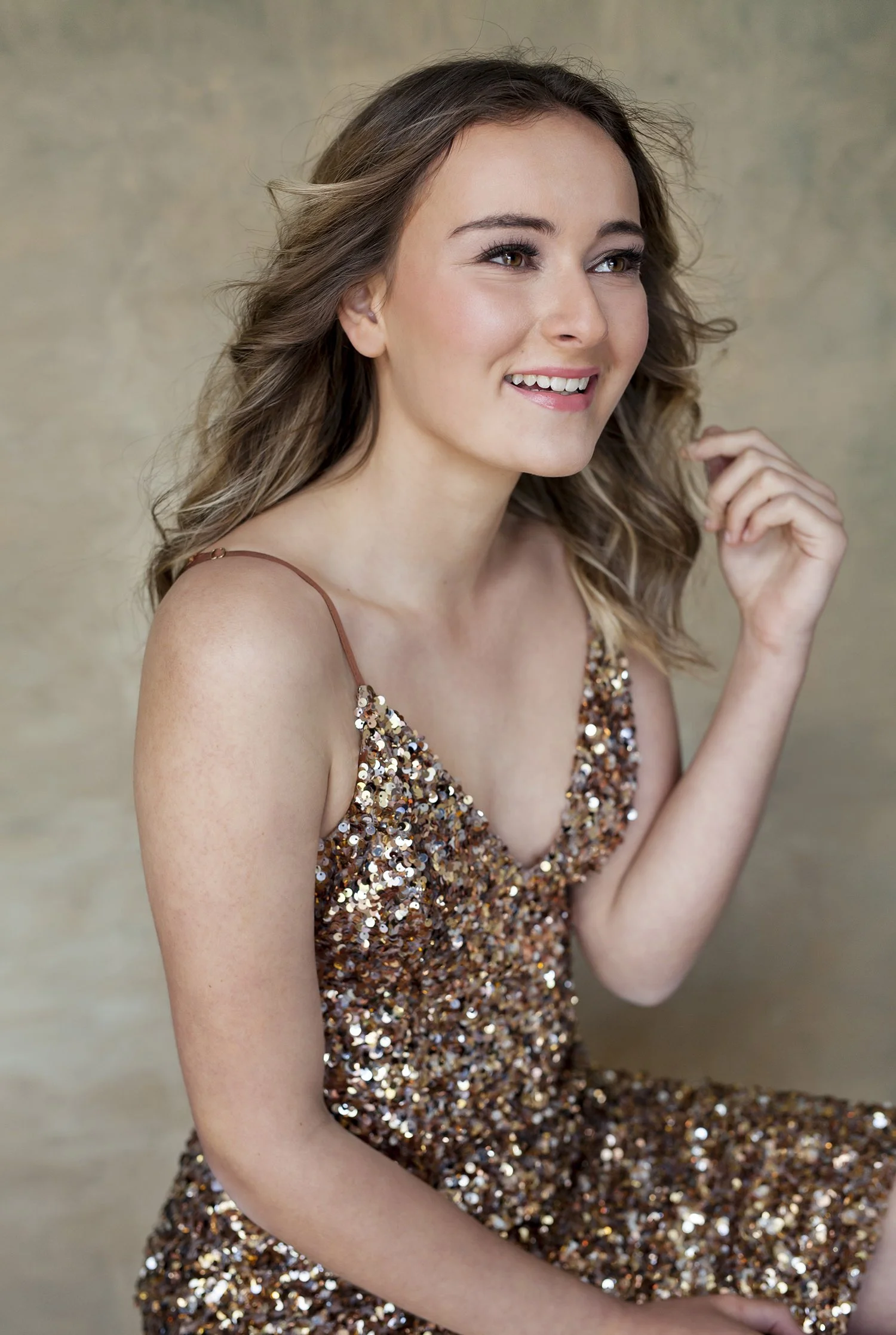 A young woman with wavy brown hair wearing a sleeveless, sequined bronze dress, smiling and looking to her left with a neutral background.