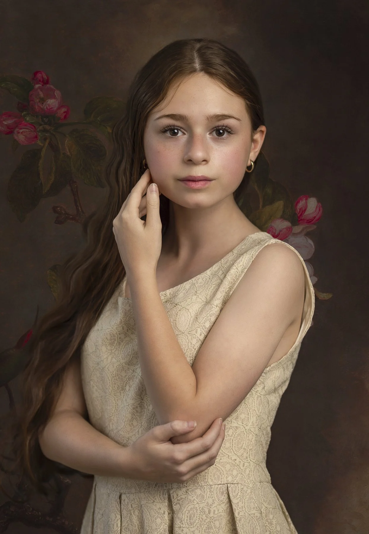 Portrait of a young girl with long wavy brown hair, wearing a sleeveless beige lace dress, standing against a dark background featuring pink flowers and green leaves.
