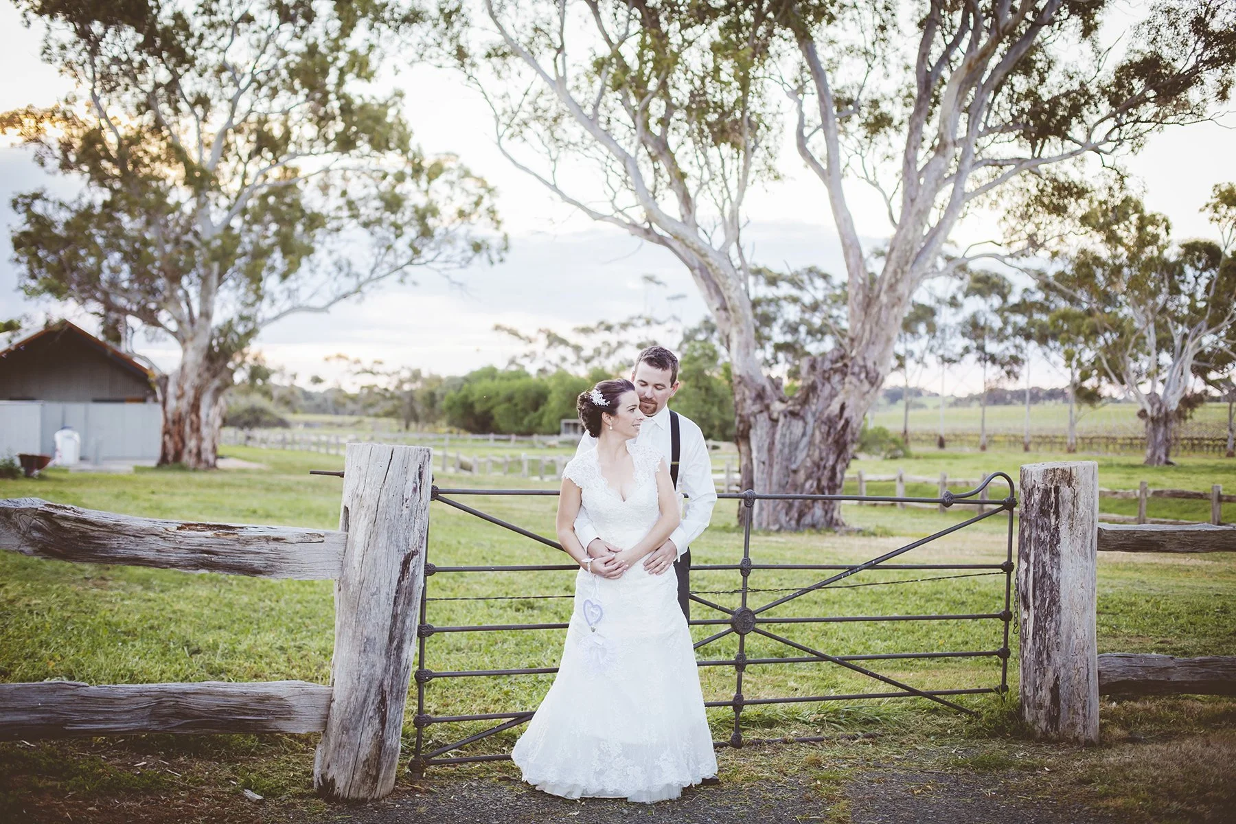 A bride and groom standing together outdoors near a rustic wooden and metal gate, with large trees and open fields in the background, during a wedding photo at sunset.
