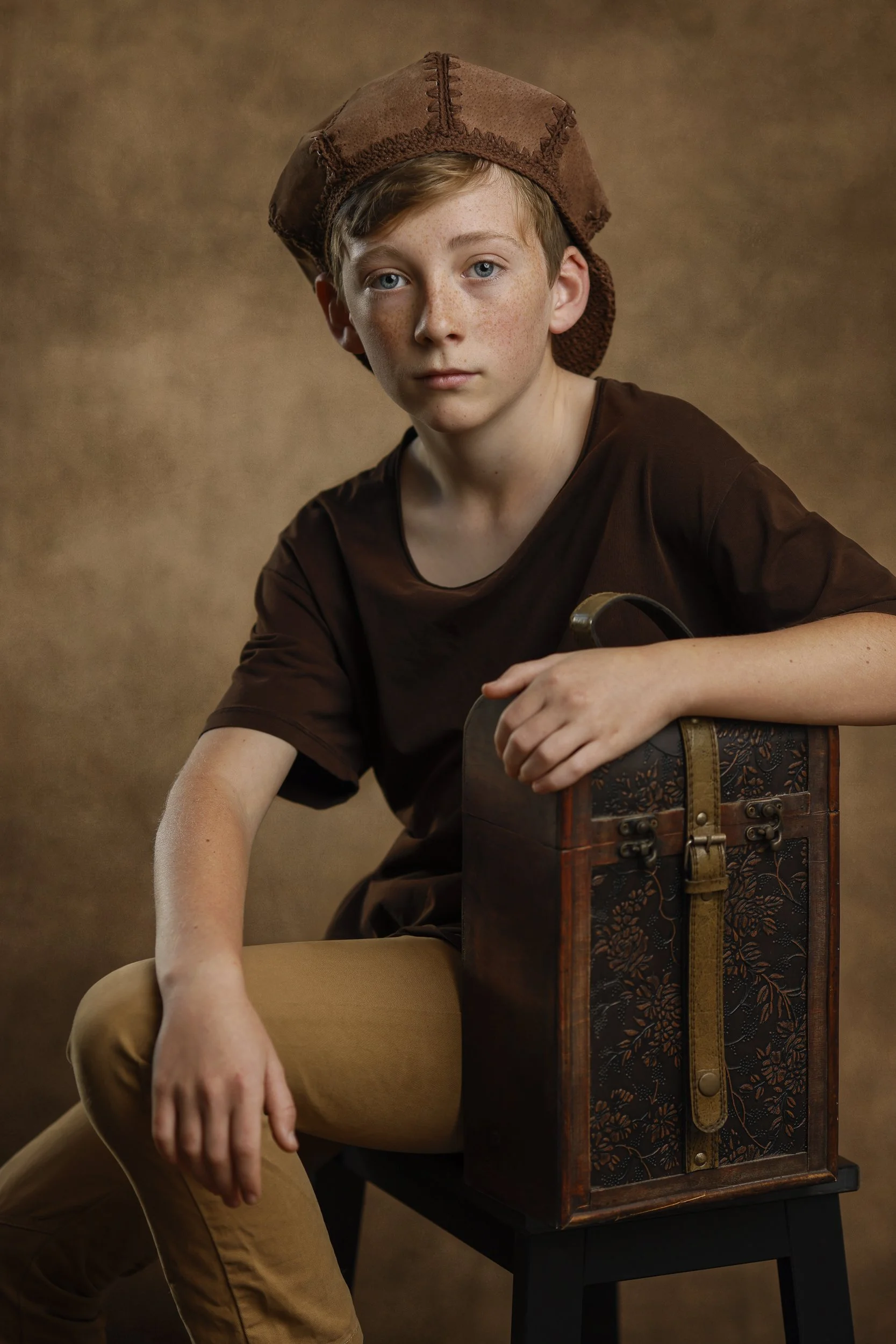 A young boy with blue eyes and freckles, wearing a brown cap and matching brown shirt, sitting on a black stool with a vintage wood and floral-patterned box.