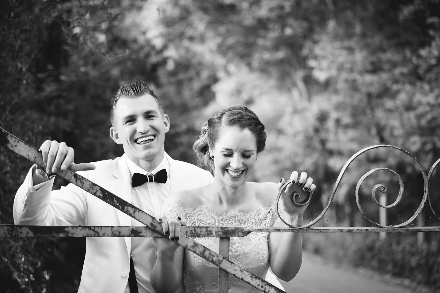 A happy bride and groom in wedding attire laughing and smiling while leaning on an ornate wrought iron gate outdoors, blurred trees in the background.