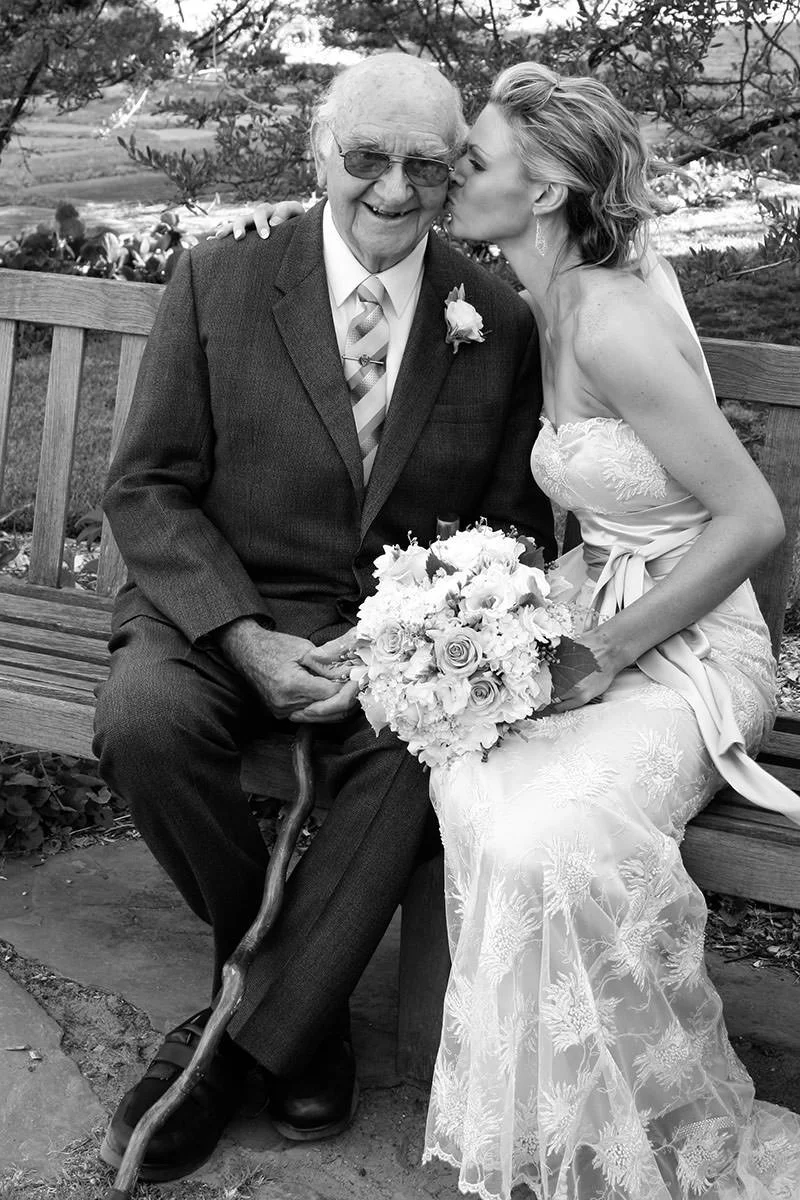 A bride in a wedding dress kisses an elderly man in a suit and tie on the cheek while seated on a bench outdoors, holding a bouquet of flowers. The elderly man has eyeglasses, a boutonniere, and a cane. There are trees and a garden in the background.