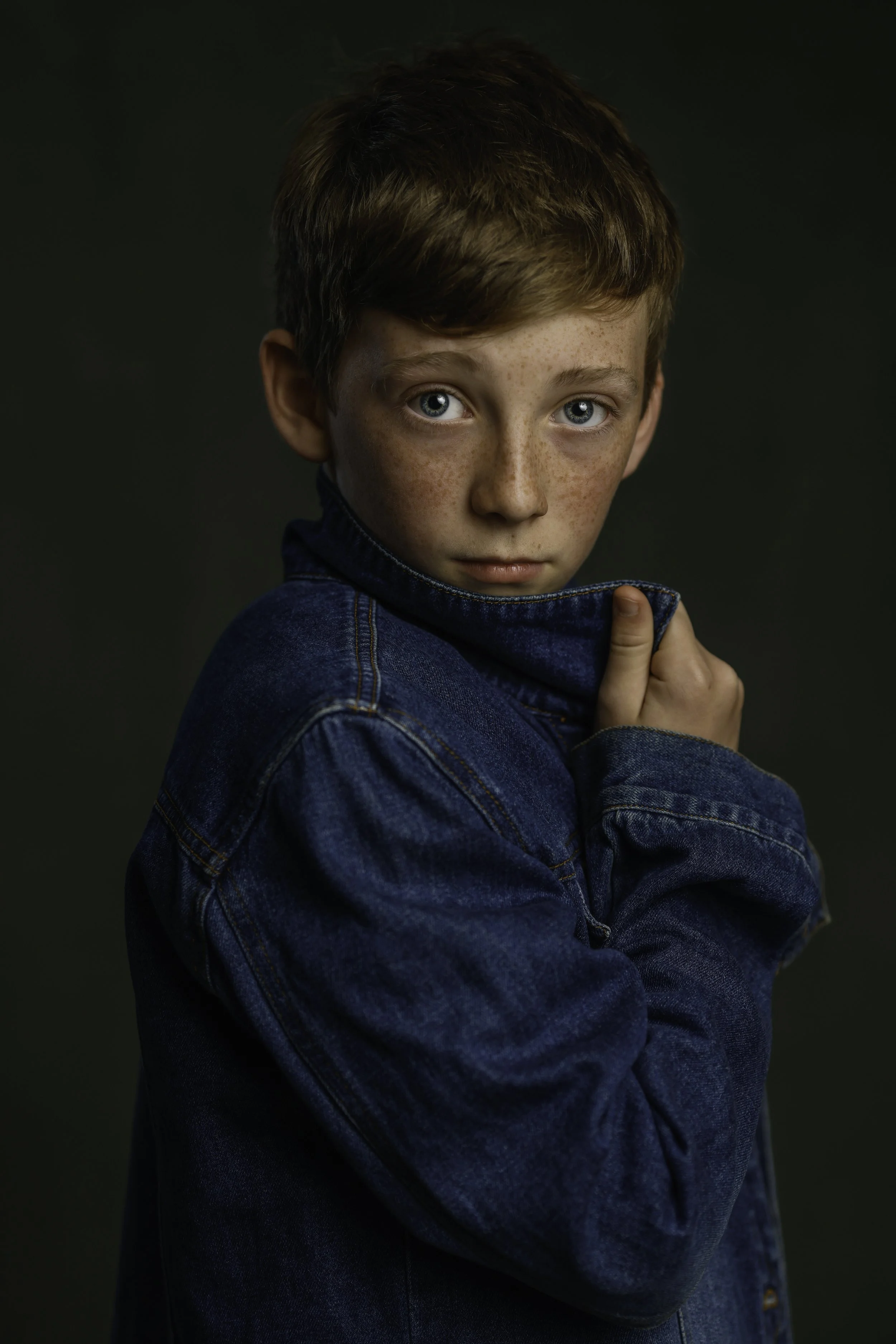 A young boy with freckles and blue eyes wearing a denim jacket, pulling the collar up to his chin, looking directly at the camera against a dark background.