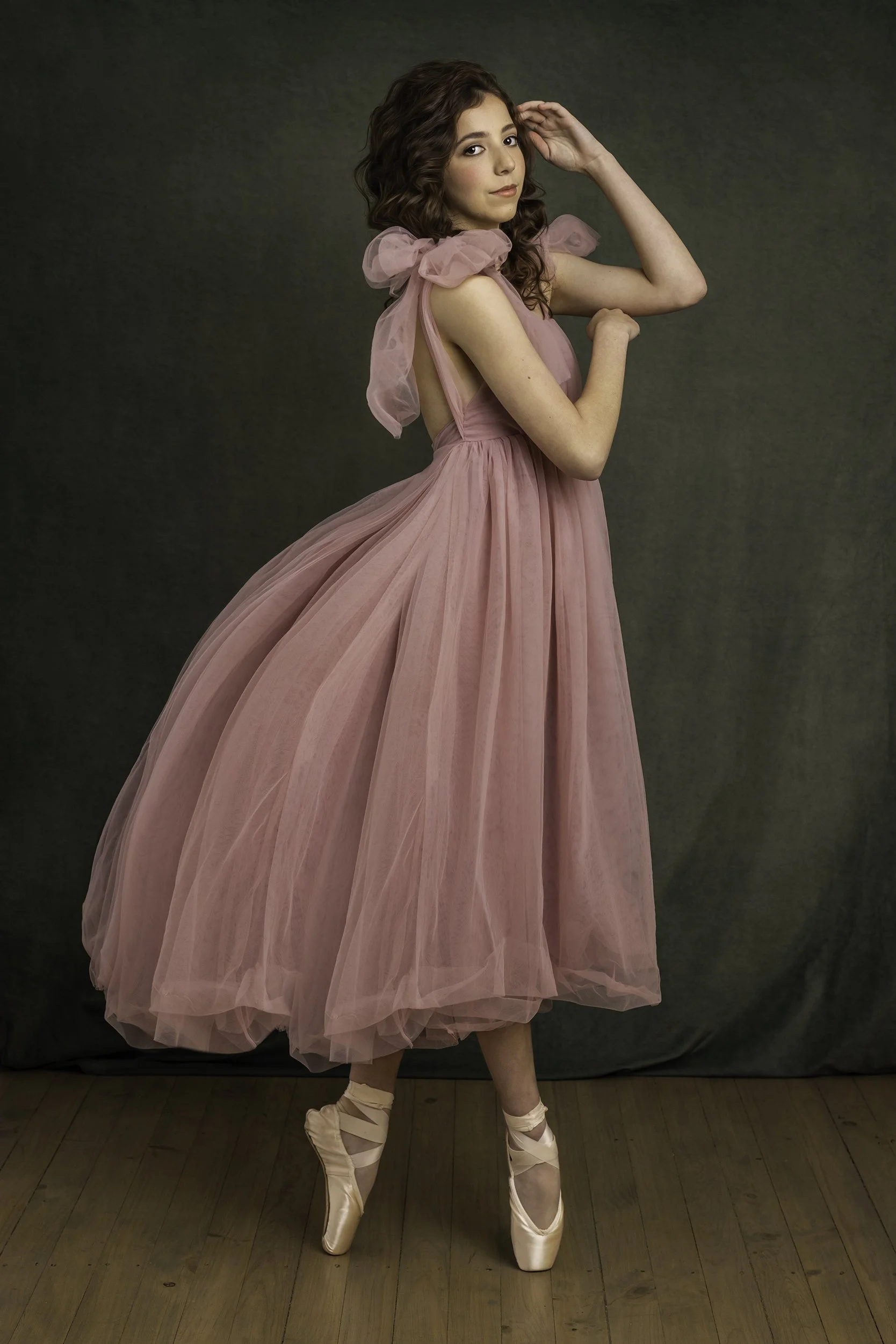 A young woman with curly brown hair in a pink, tulle, ballet-inspired gown standing on pointe shoes against a dark neutral background.