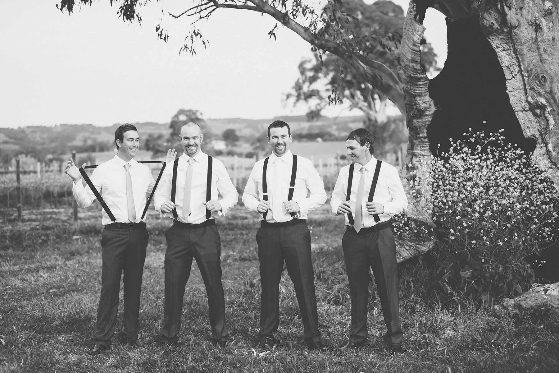 Four men in formal attire standing outdoors, smiling and laughing, with large trees and open land in the background.