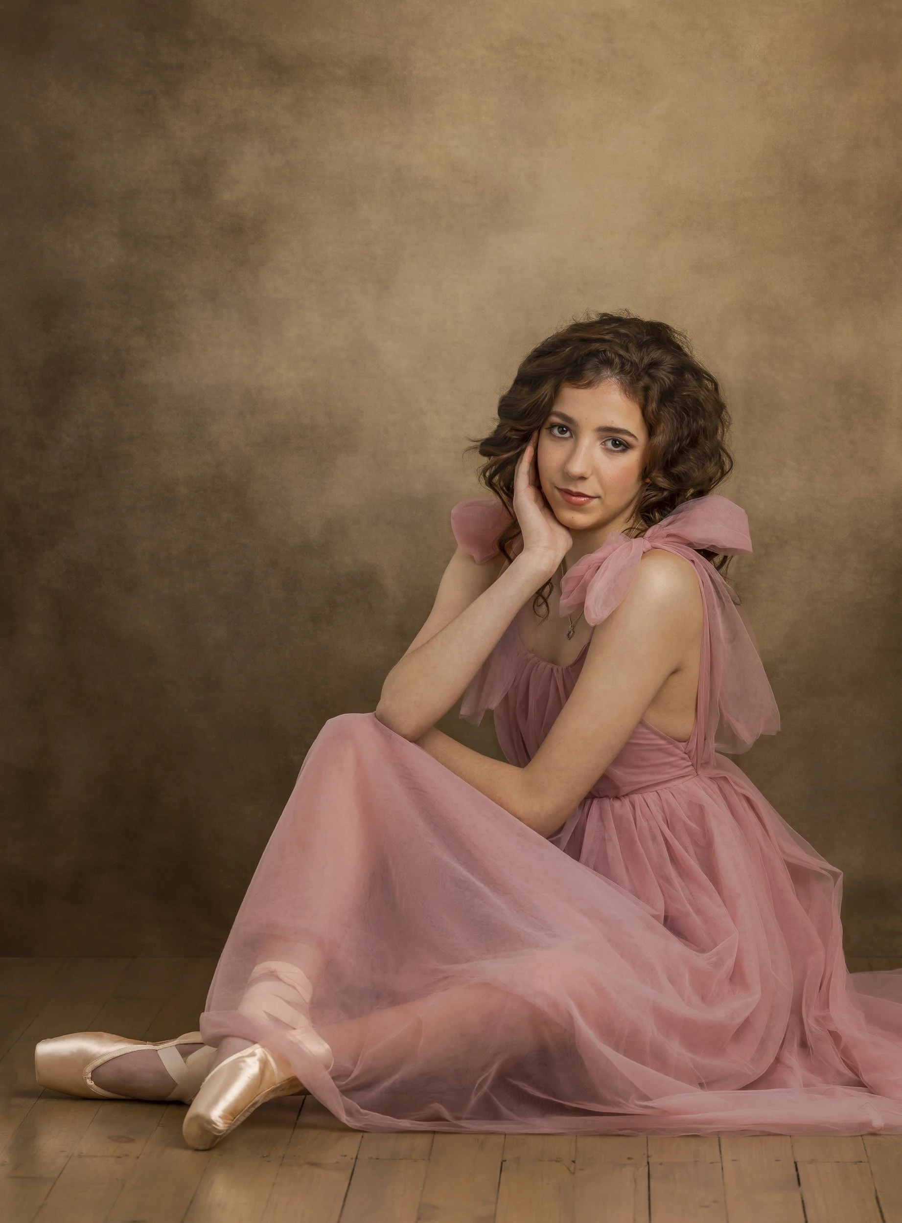 A young woman with curly brown hair wearing a pink ballet dress with satin pointe shoes sitting on a wooden floor and posing in front of a brown textured background.