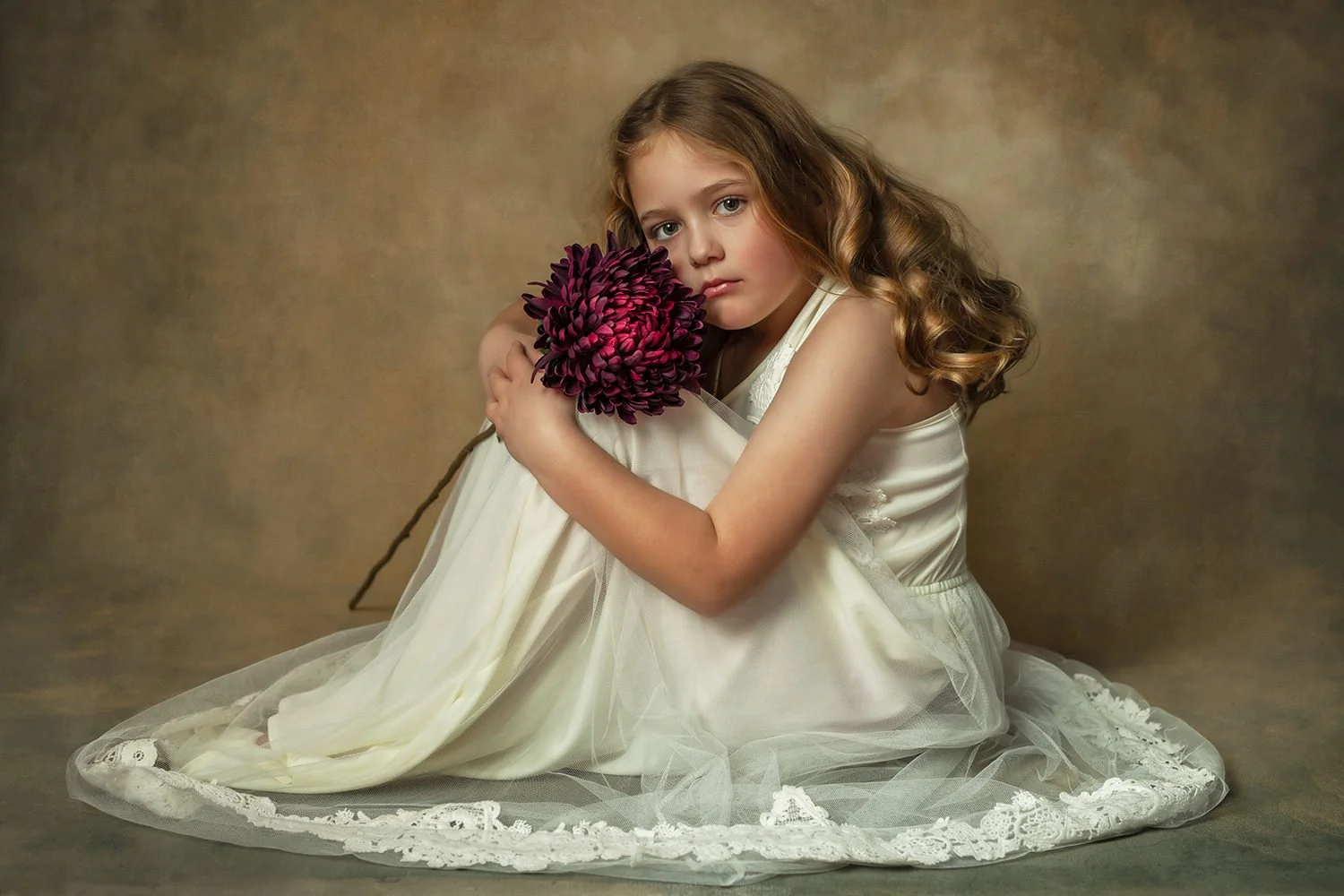 A young girl with long, curly brown hair sitting on the floor in a white dress, holding a large purple flower-shaped object close to her face, with a contemplative expression, against a neutral brown background.