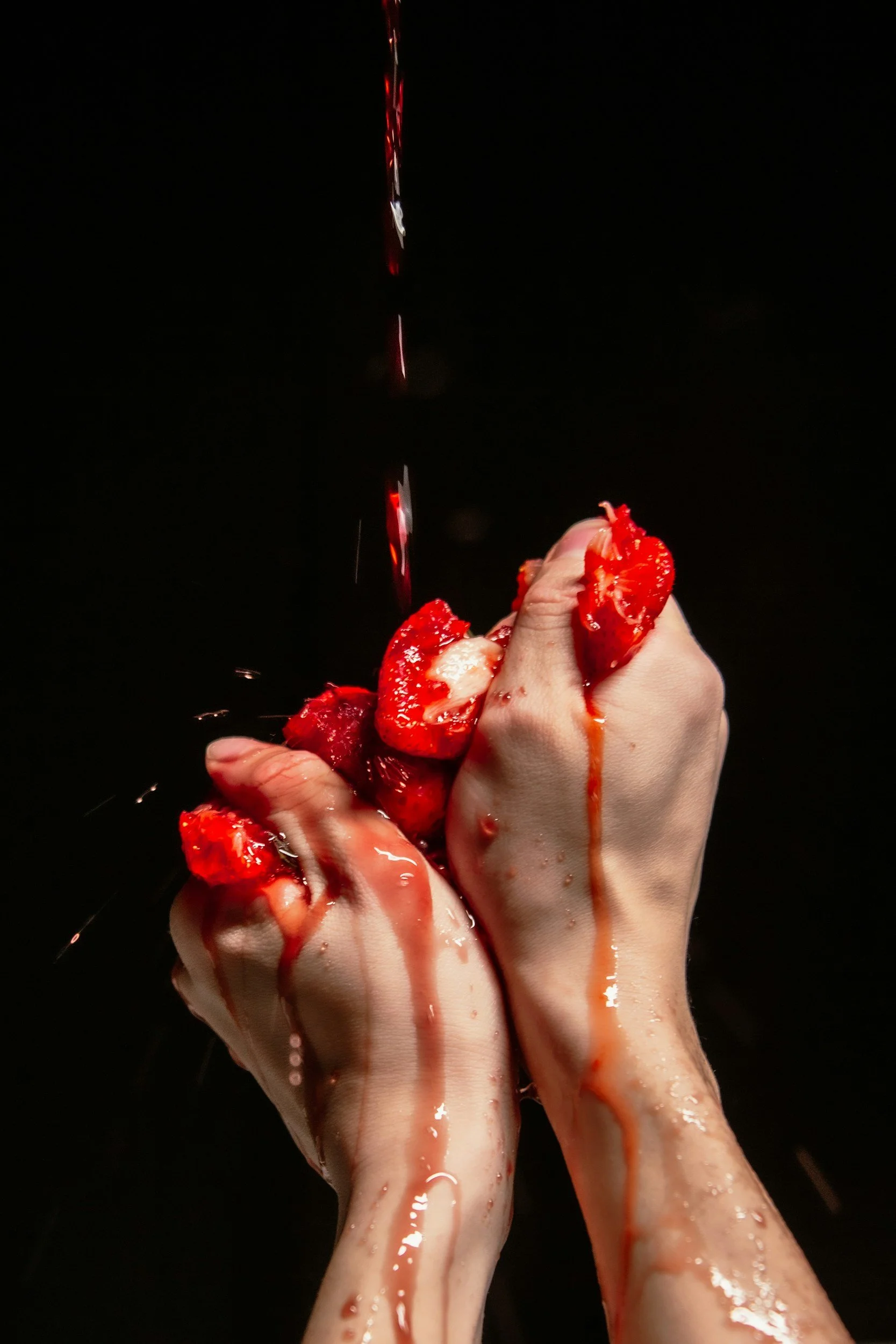 Two hands clenched in a fist covered in sliced strawberries and strawberry juice, with liquid dripping down onto a black background.