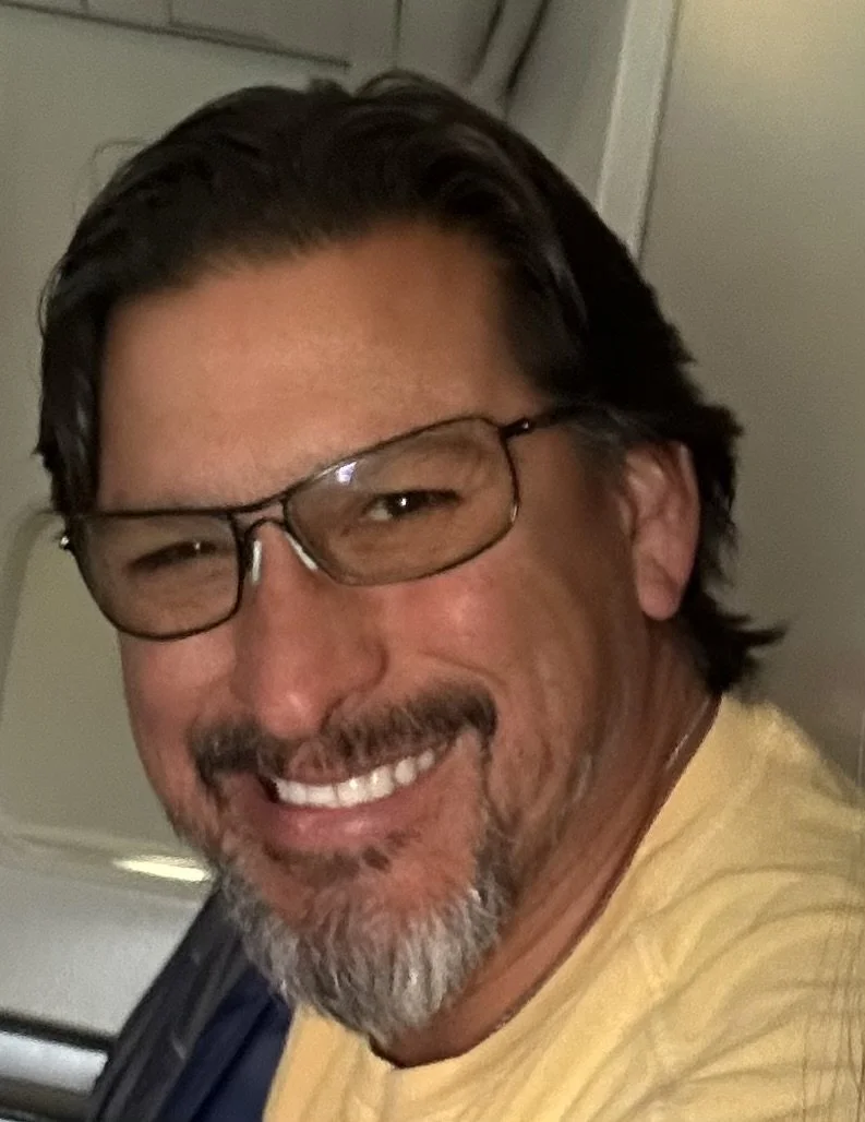Close-up of a smiling man with dark hair, glasses, a beard, and wearing a yellow shirt.