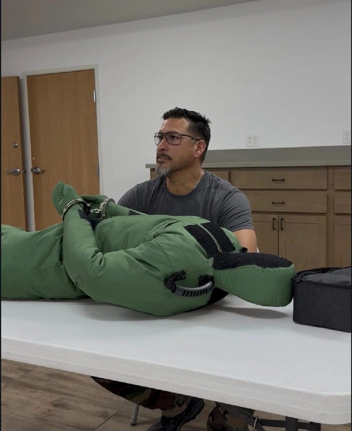 A man with glasses and a beard sitting at a table with a person in green protective gear lying down.