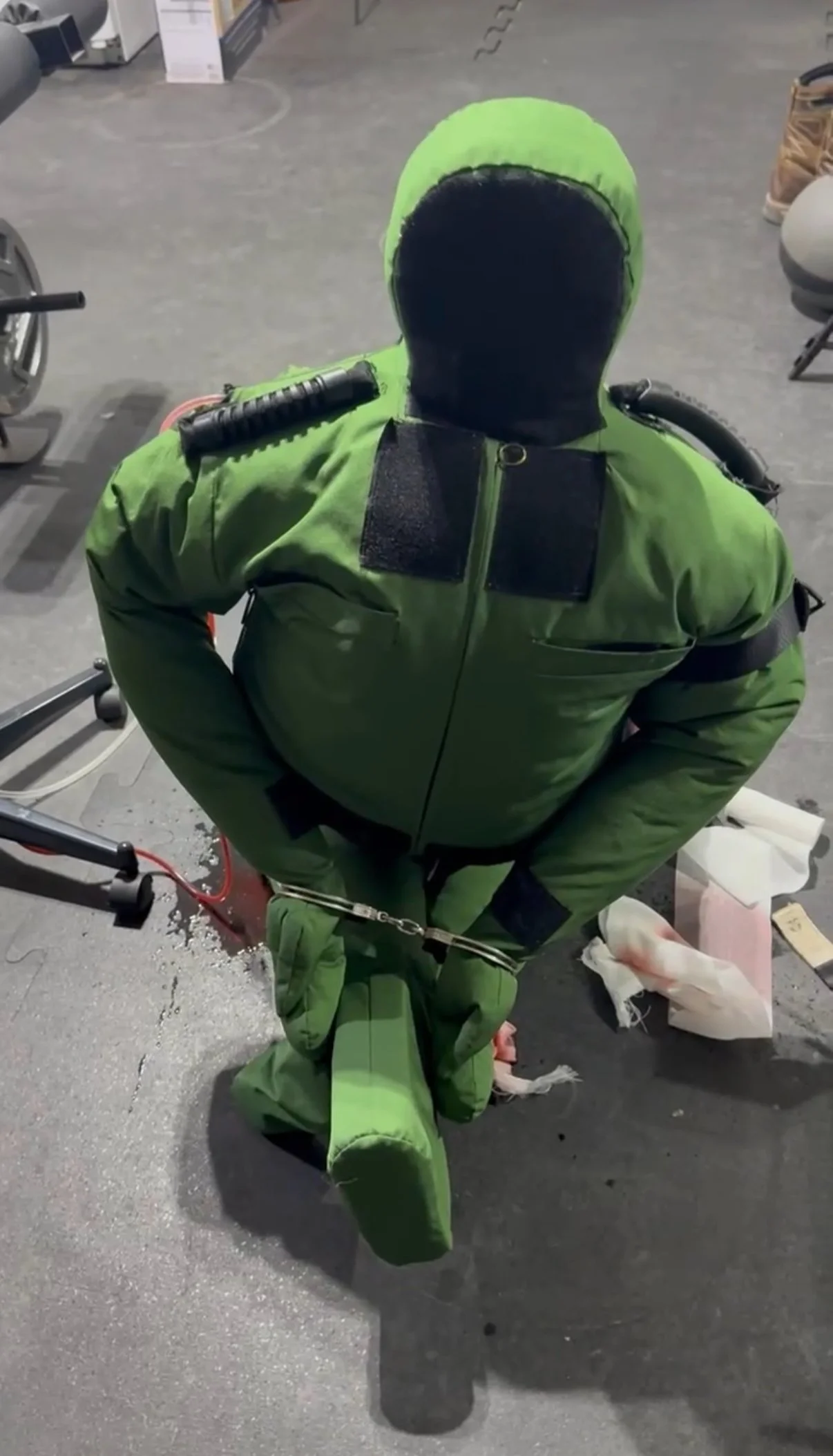 A person in a green protective suit and mask kneeling on the floor, restrained with handcuffs, in a room with various equipment and supplies.
