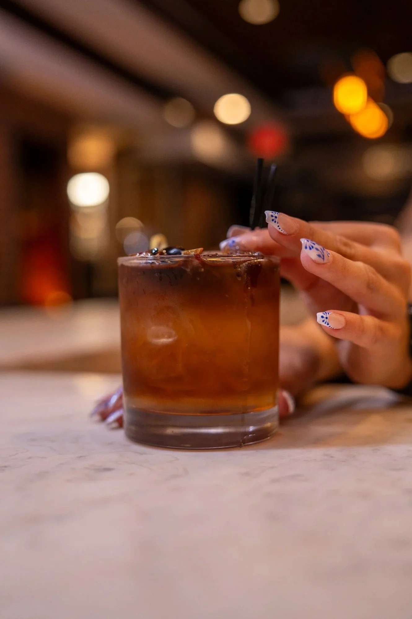 A person’s hand with decorated nails holding a glass of iced coffee or cocktail with a dark liquid and ice, on a marble countertop in a dimly lit bar or restaurant setting.
