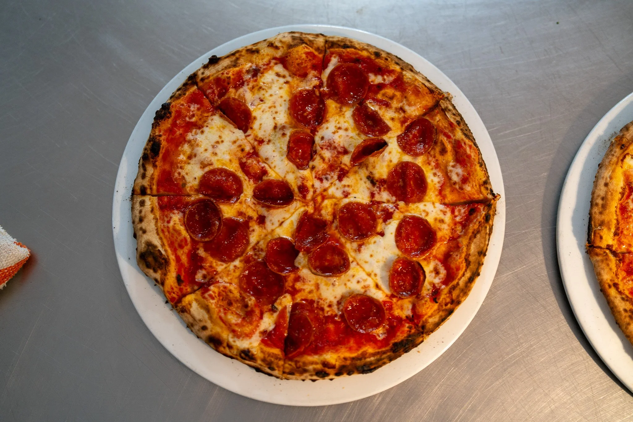 Pepperoni pizza on a white plate with a slightly charred crust, placed on a metal surface.