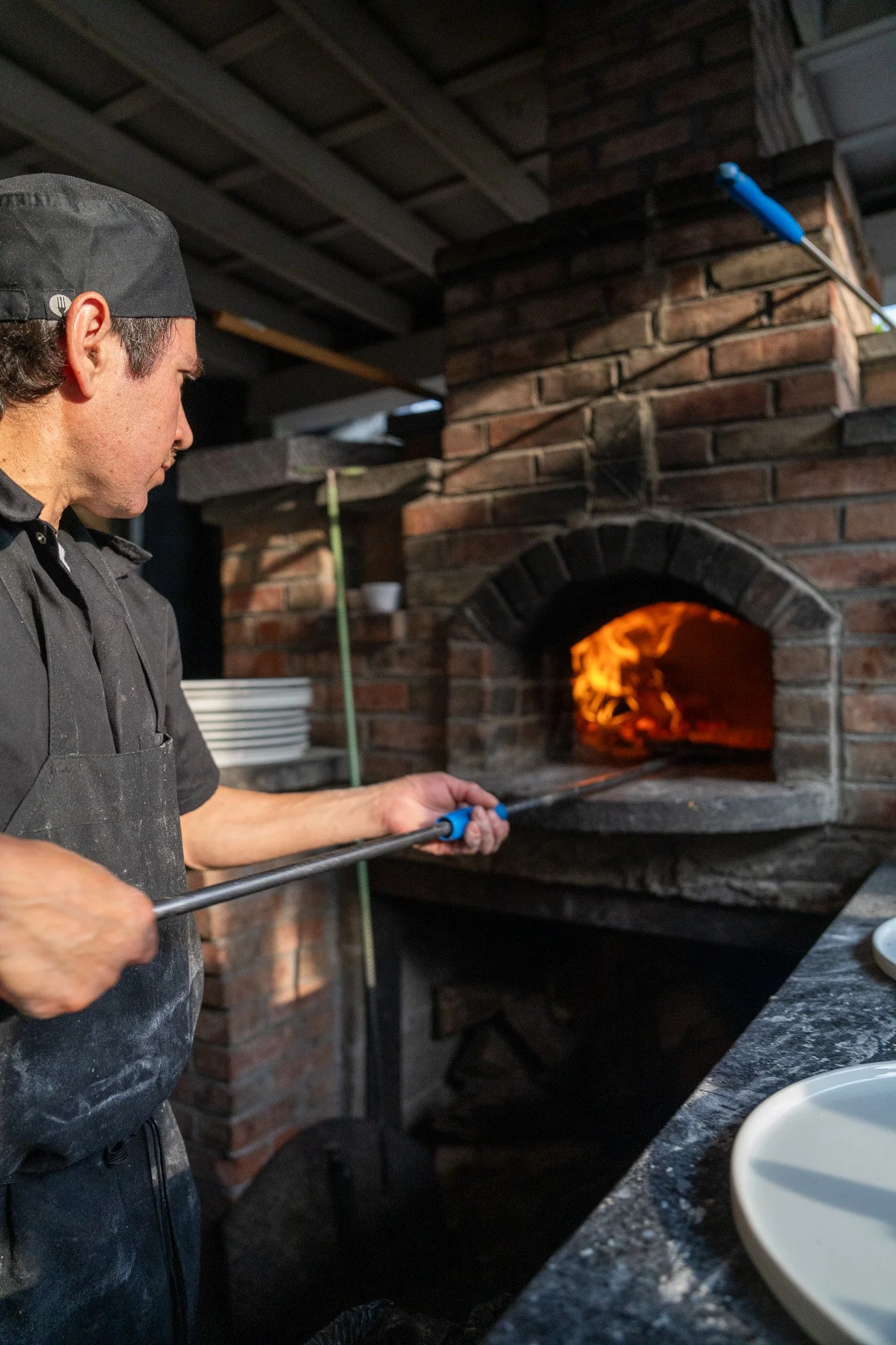 A chef using a blowtorch to sear food in front of a brick oven with a visible flame inside.
