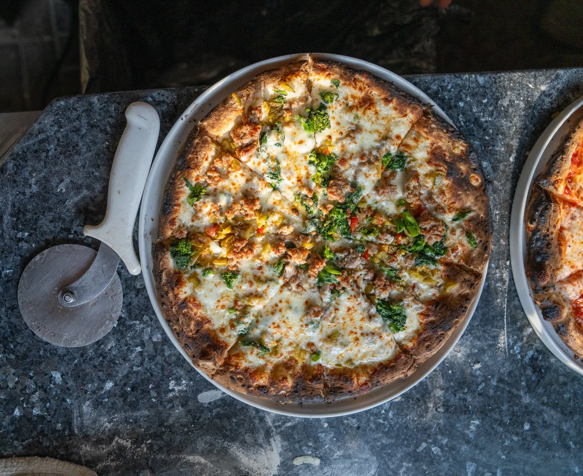 A pizza with cheese, green vegetables, and ground meat on a round metal tray, placed on a dark granite surface.