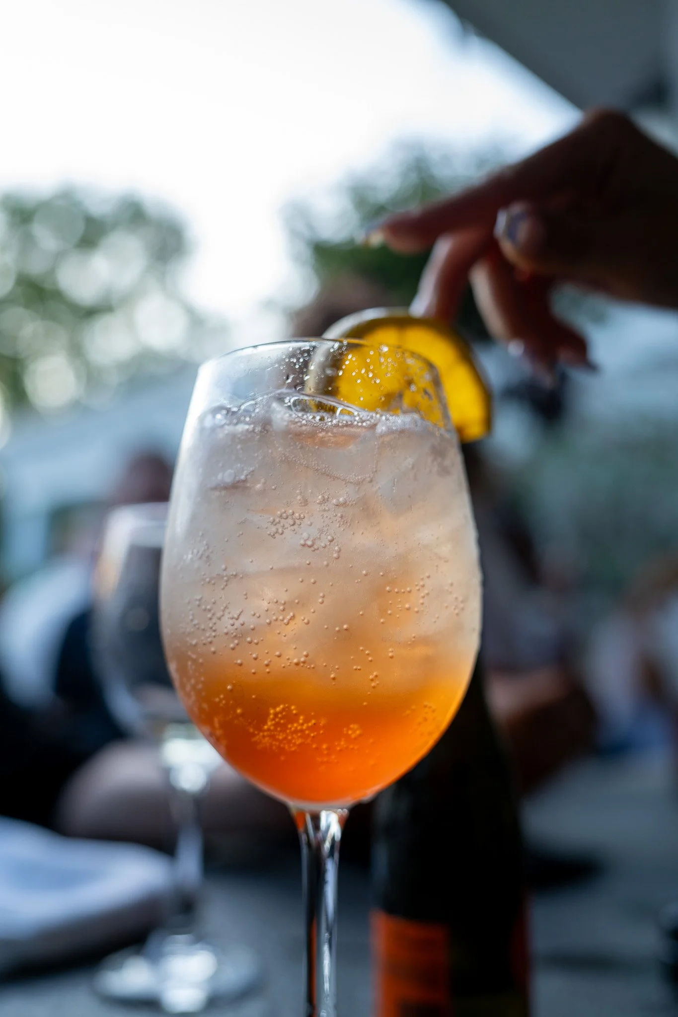 A person's hand holding a glass with a lemon wedge, filled with a bubbly, pinkish cocktail, with a blurred background.