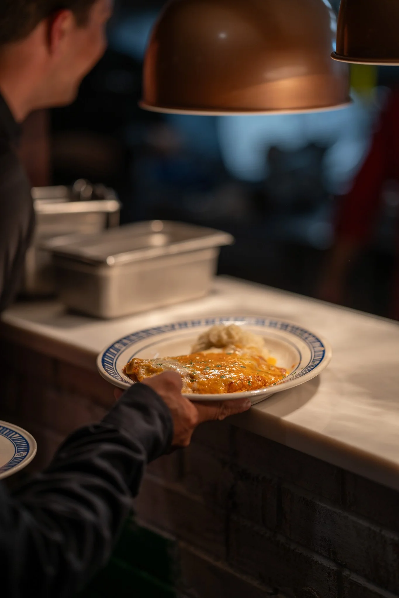 Person serving plate of food, possibly lasagna, with mashed potatoes on the side at a restaurant or cafeteria.