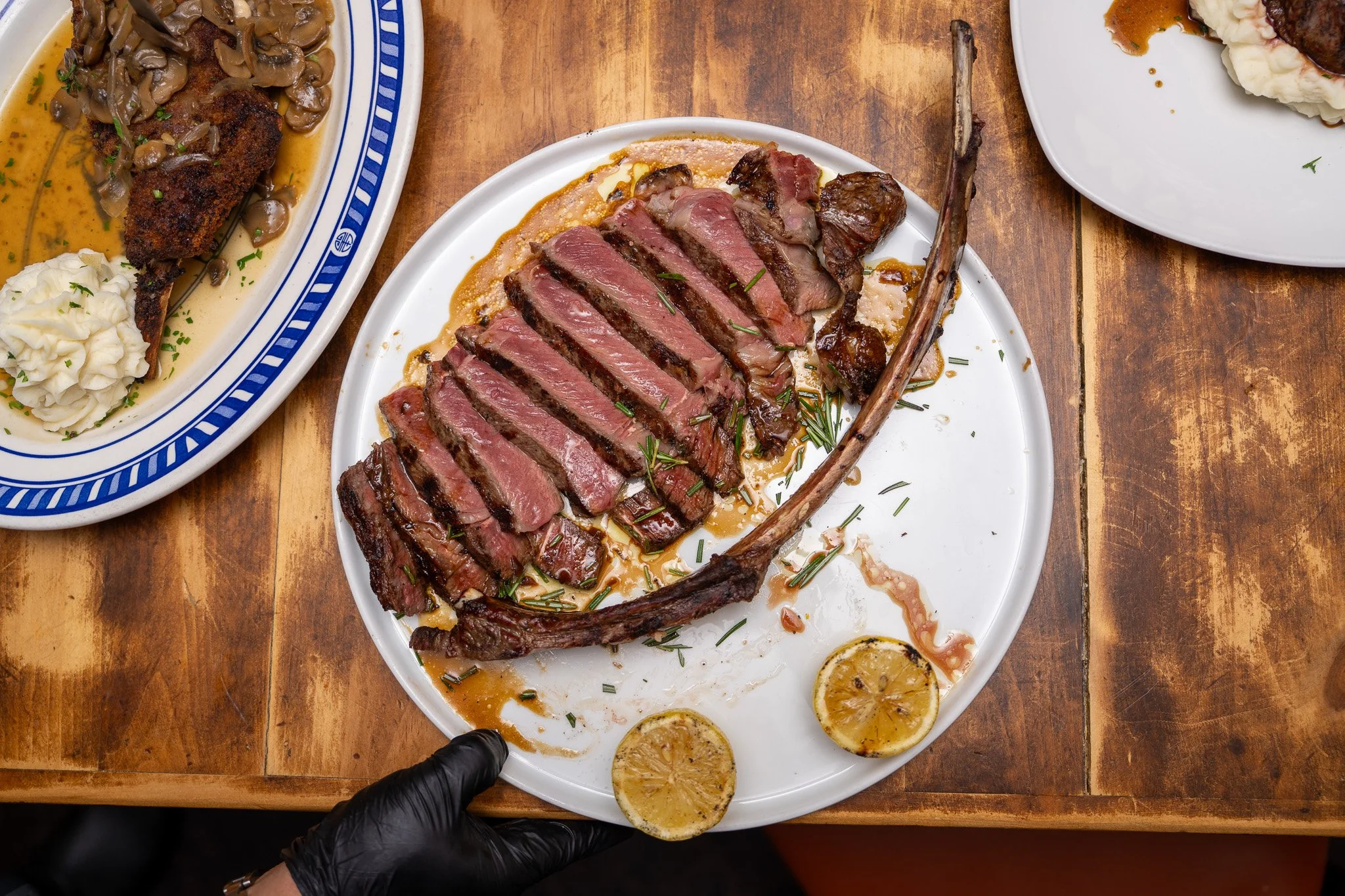 A plate with sliced medium-rare steak garnished with chopped herbs, lemon slices, and sauce, on a wooden table.