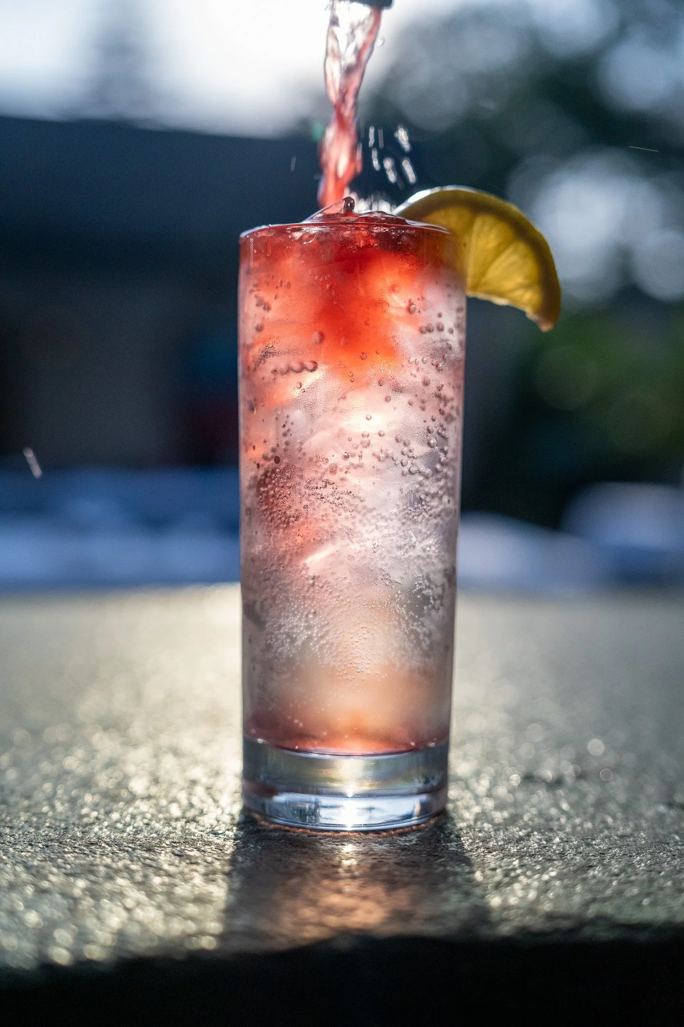 A tall glass of sparkling pink lemonade with a lemon wedge on the rim, ice cubes, and bubbles, being filled with water.