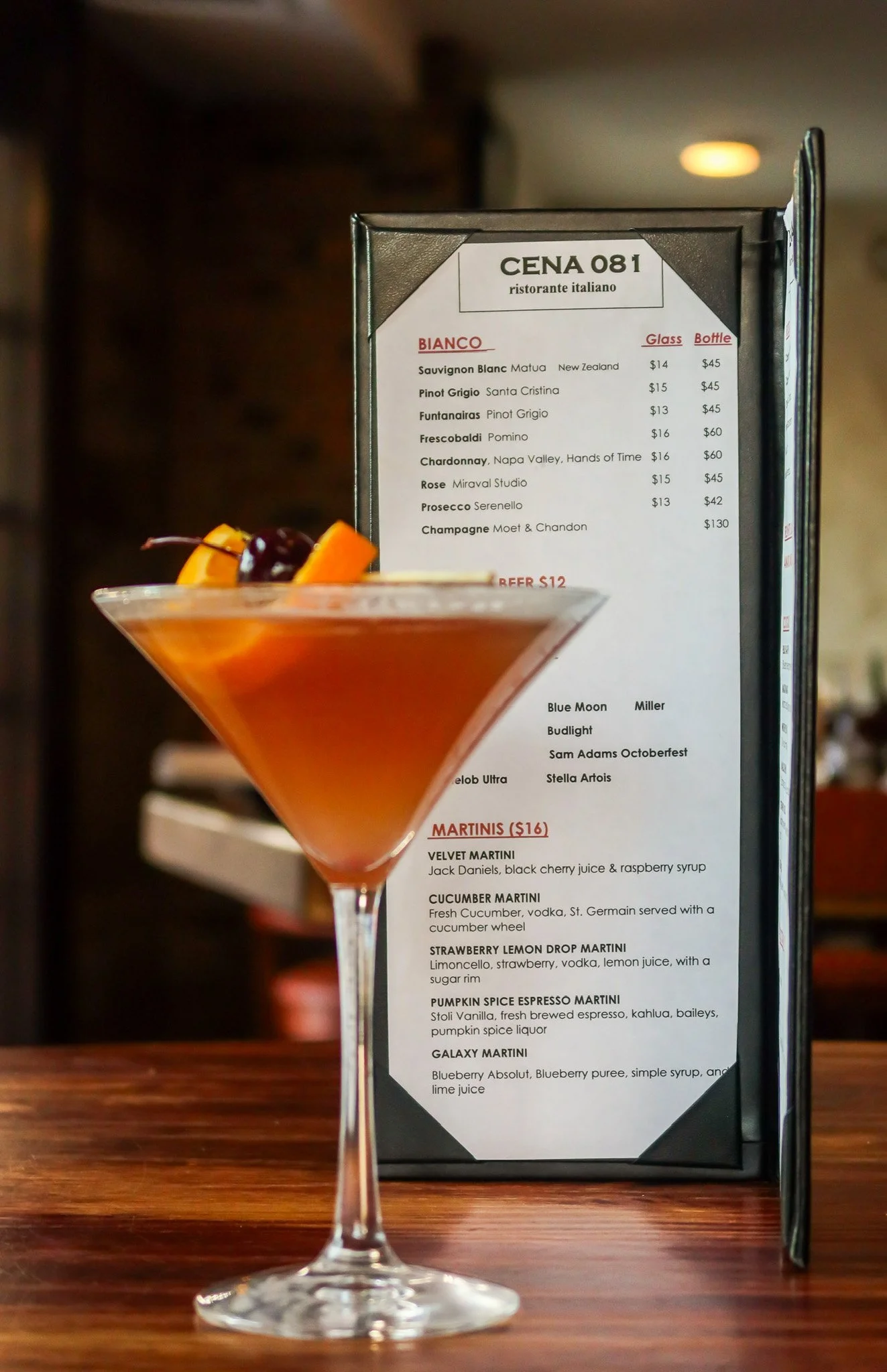 A cocktail in a martini glass garnished with orange slices and a cherry in front of a restaurant menu.