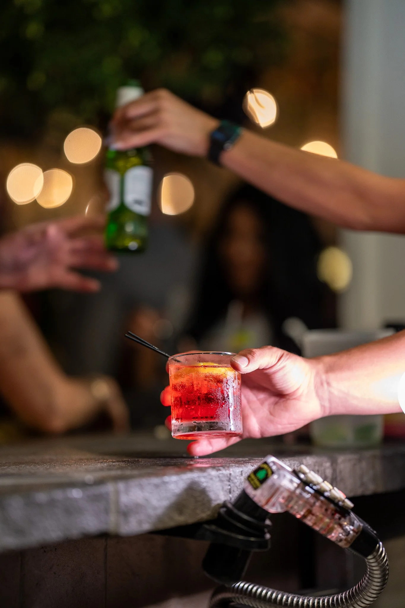 A bartender hand holding a red cocktail with a straw, with glasses of beer and drinks in the background in a bar setting.