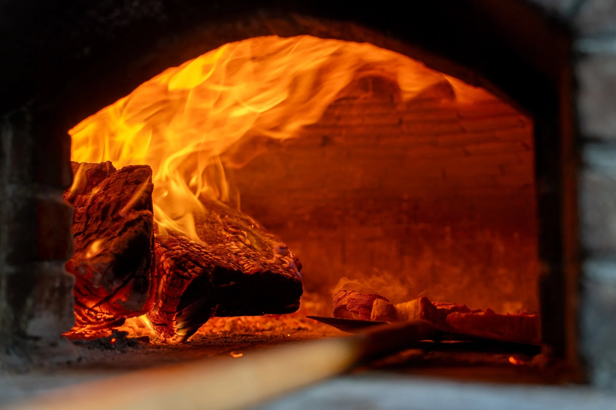 Fire burning inside a brick oven, with flames surrounding a piece of wood.