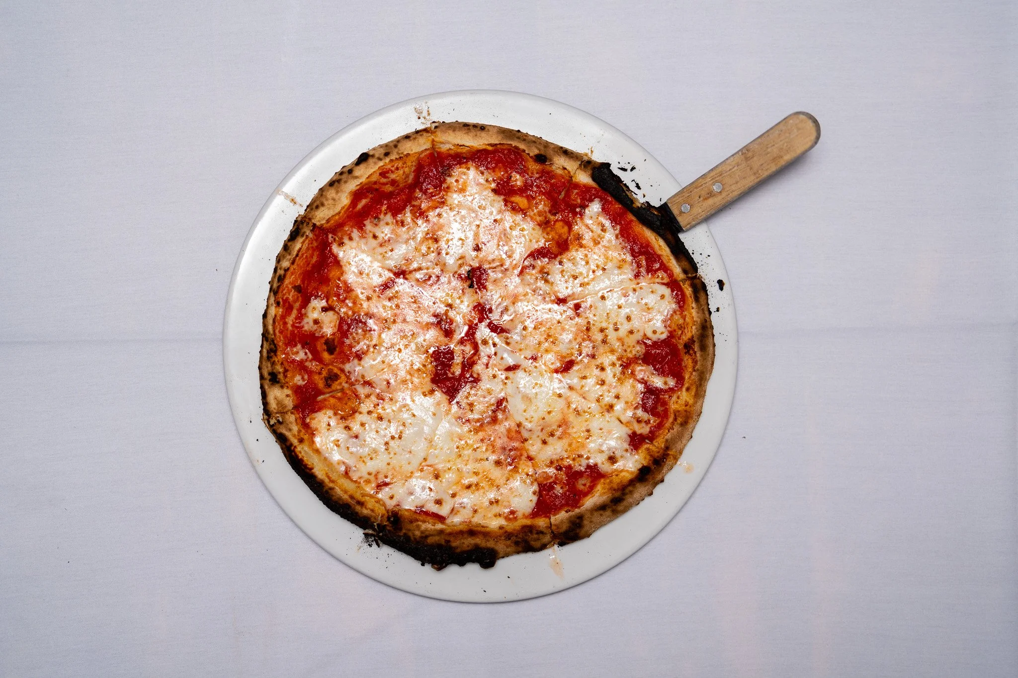 A cheese pizza on a white plate with a metal pizza cutter resting on the side.