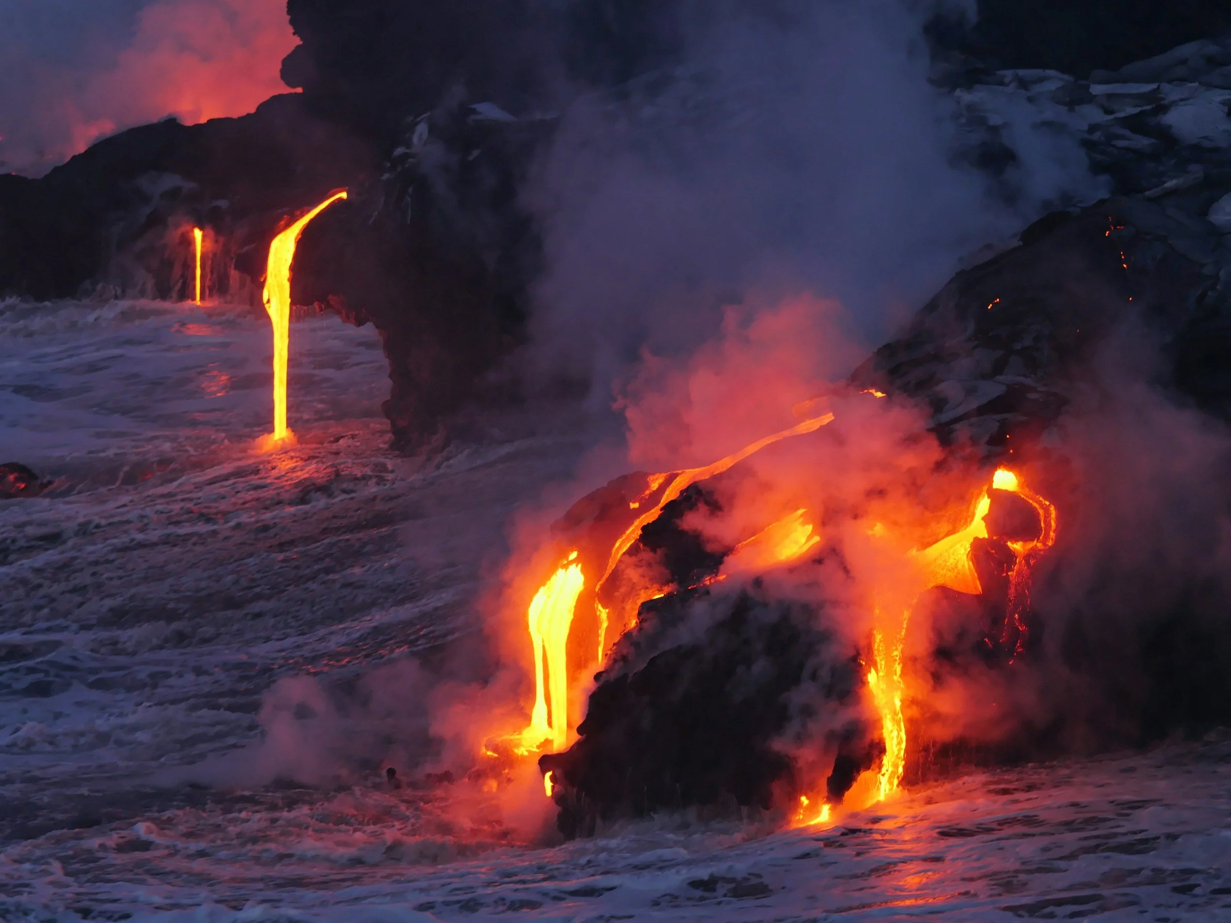 Eruption of lava flowing from volcanic rocks into the ocean, with steam and smoke rising at sunset.
