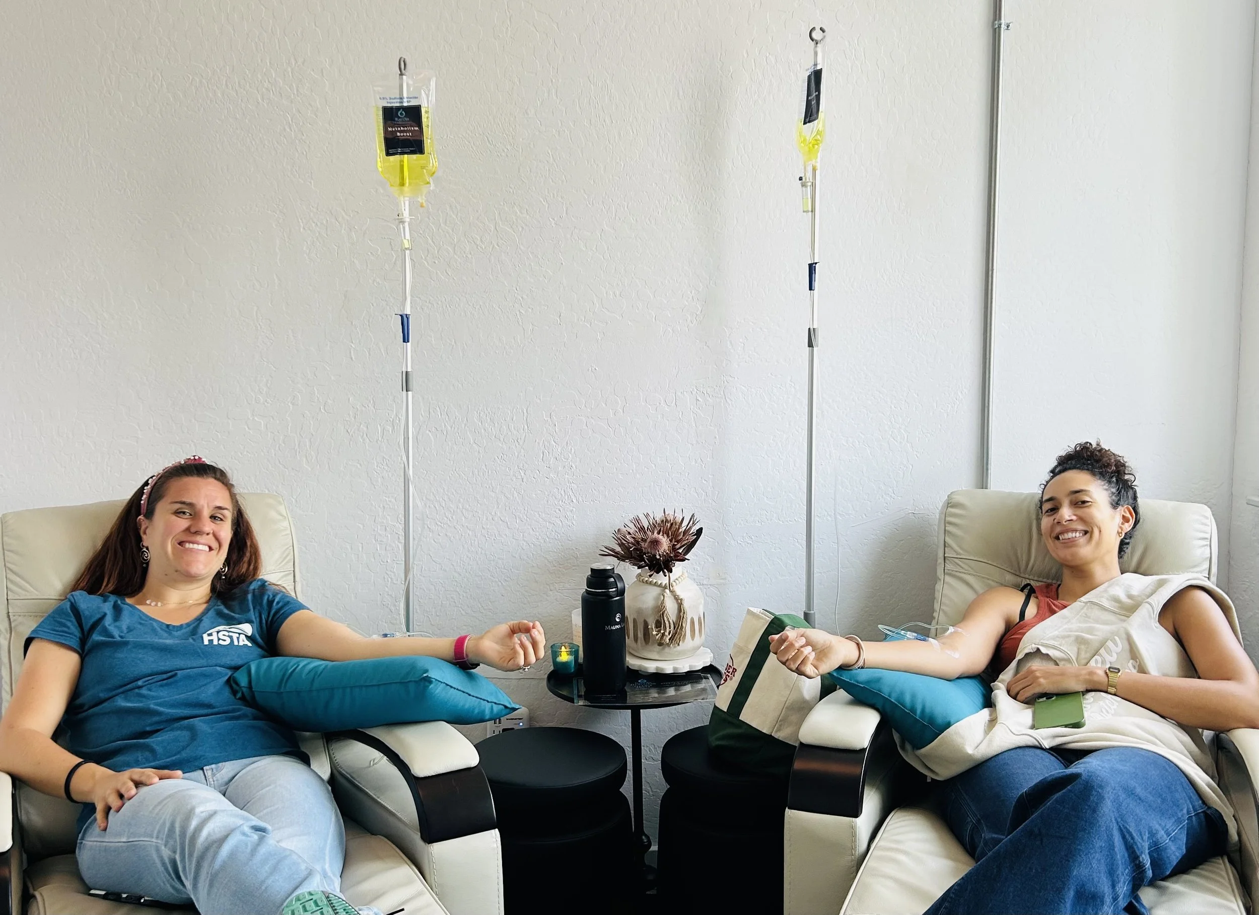 A gloved hand holding two syringes filled with a red liquid in front of green tropical plants.