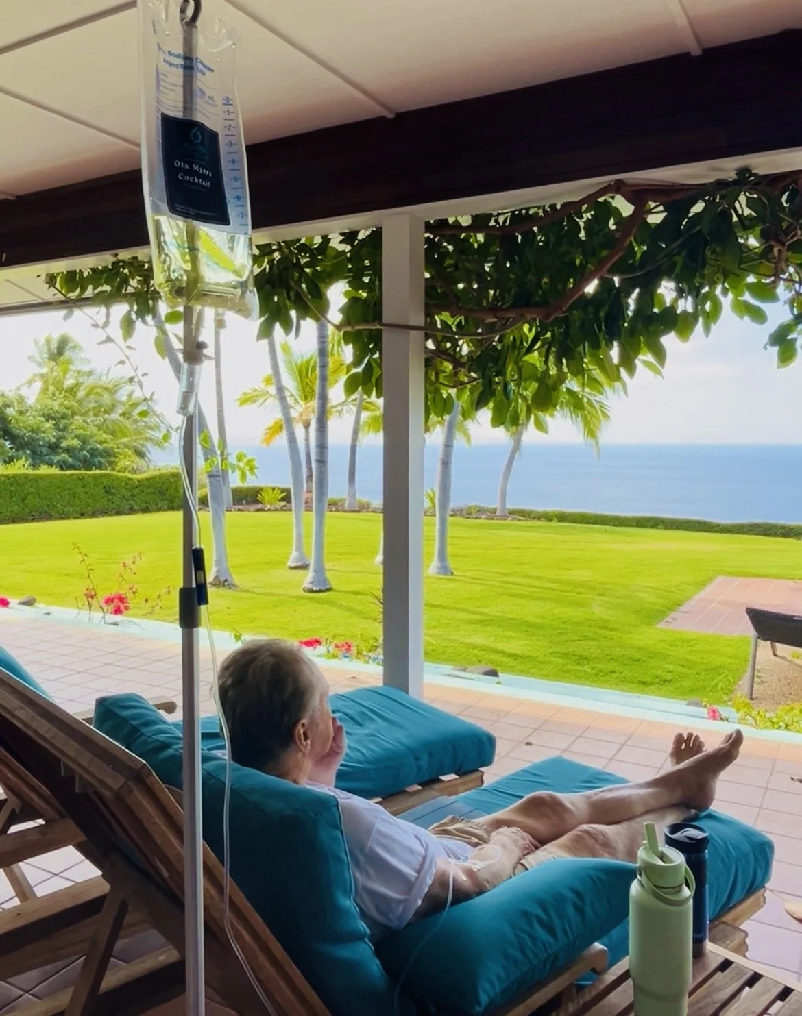 People relaxing on lounge chairs by a pool overlooking the ocean on a cloudy day, with a thatched umbrella and IV drip stands nearby.